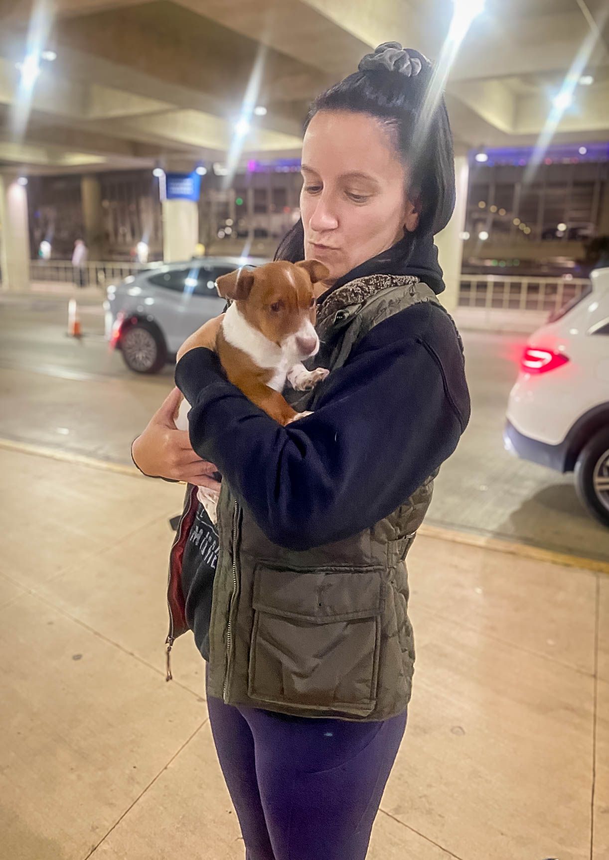 Young woman holding a small brown and white puppy outside at night near parked cars.