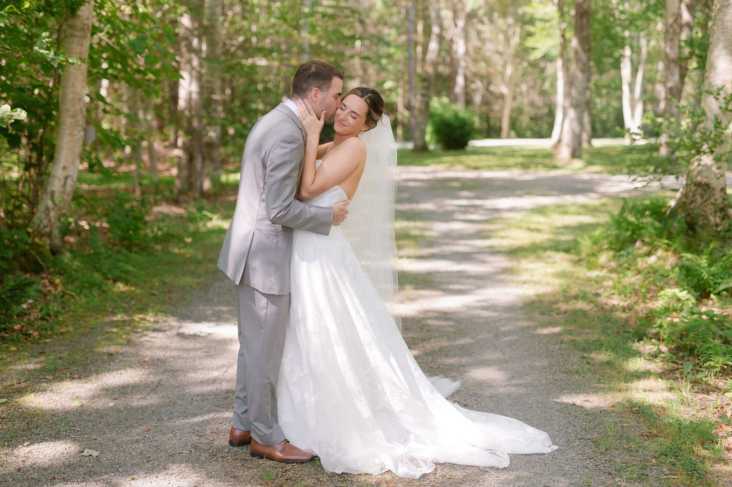 Romantic couple in formal wedding attire embraces while walking along a sunlit wooded path.