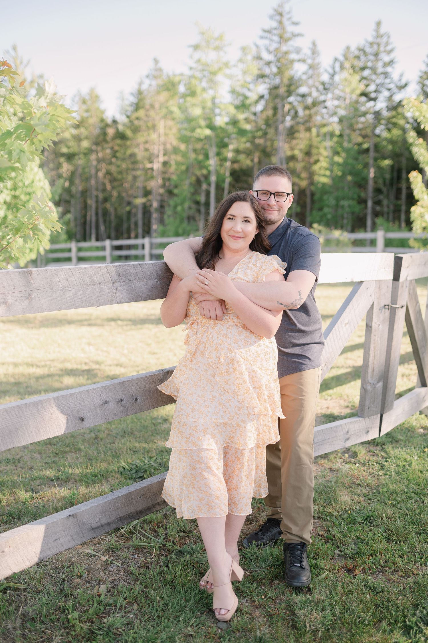 A couple poses playfully on a wooden fence in a rural setting.