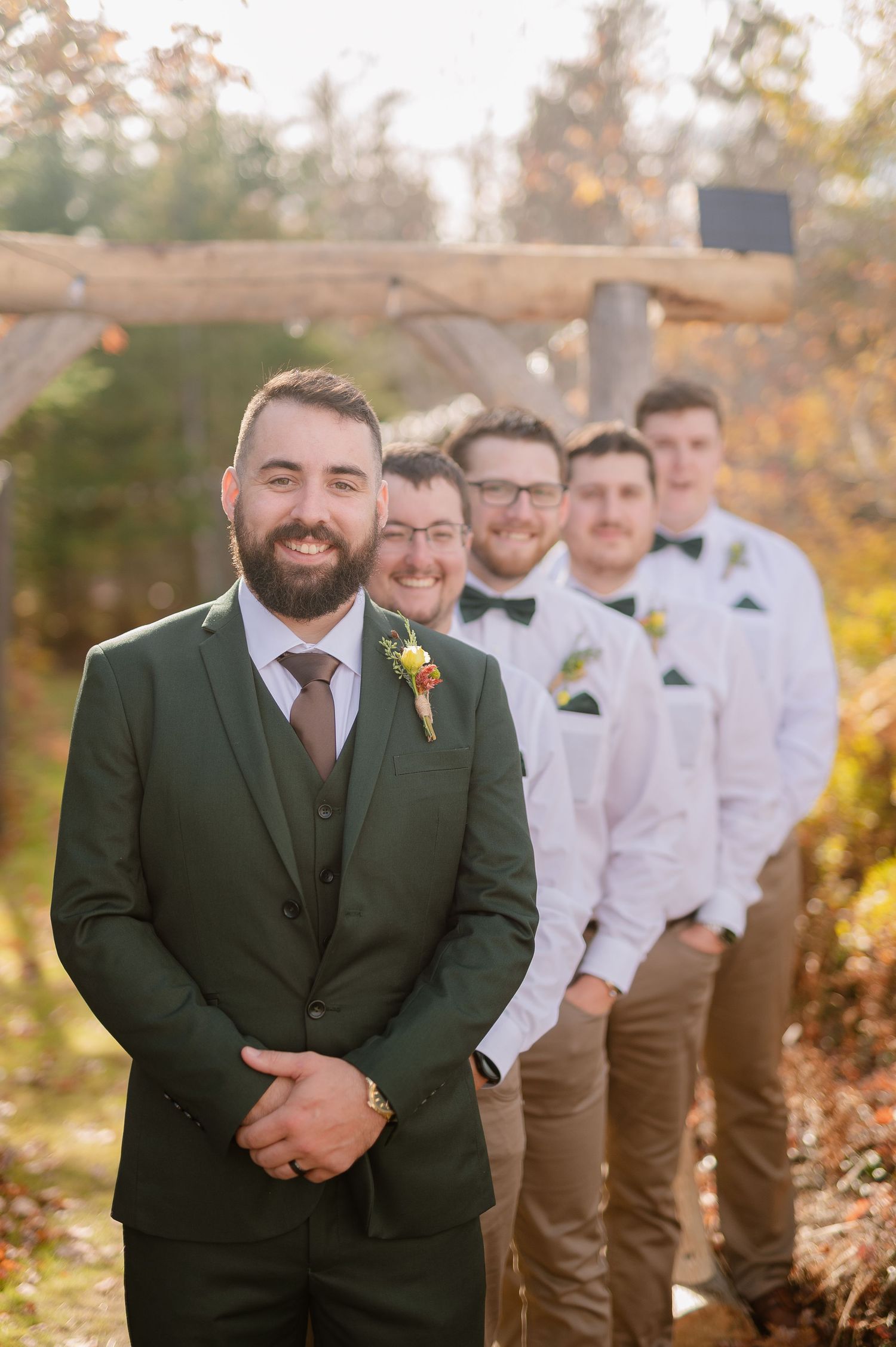 Groomsmen lined up in dark suits and white shirts against autumn foliage backdrop.