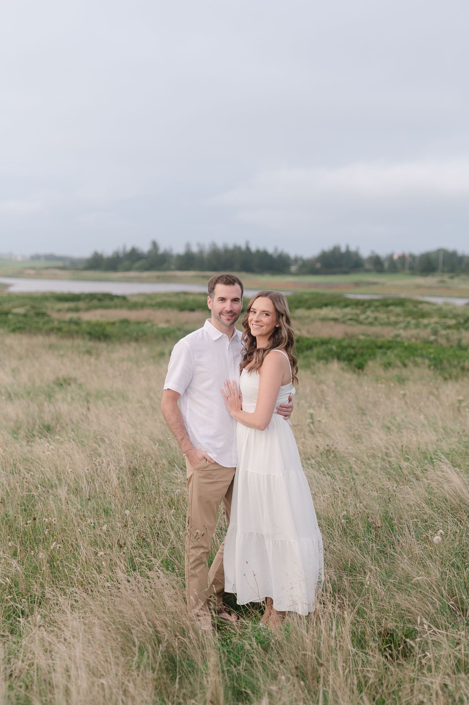 A romantic couple embraces while standing in tall grass with trees in the background.