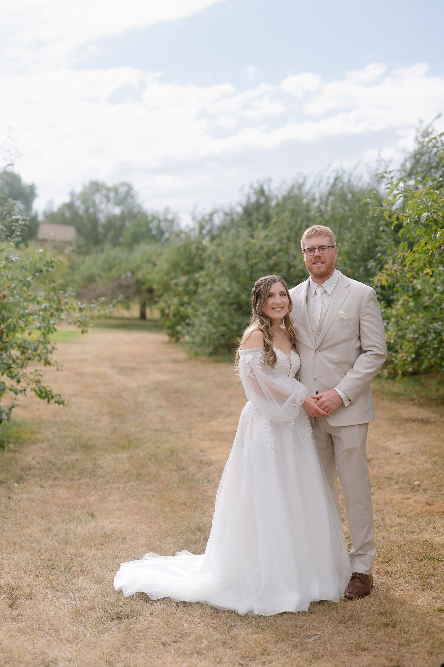 Wedding couple walks through a sunny apple orchard in formal attire.