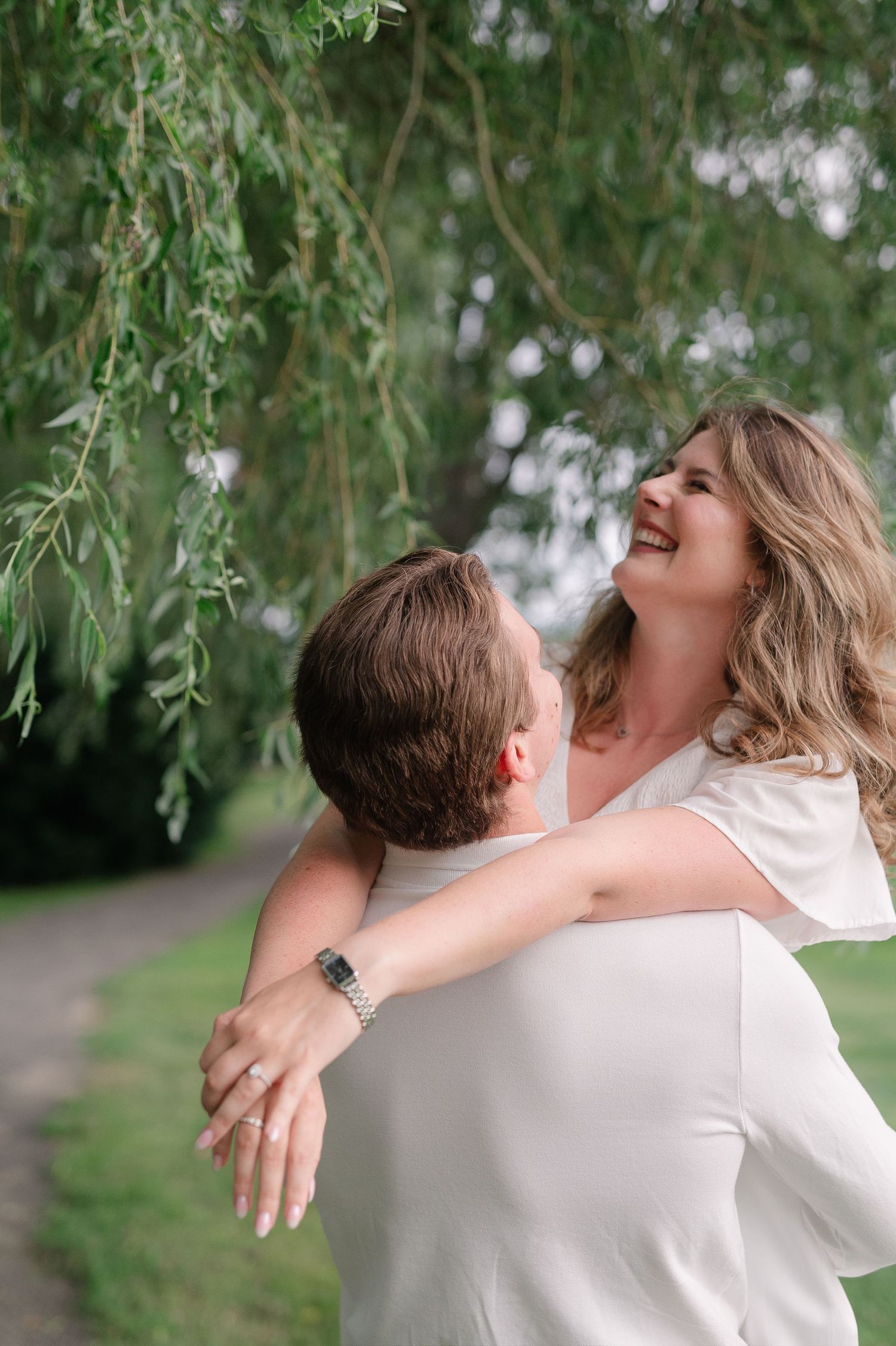 A joyful couple embraces and laughs together under drooping willow tree branches.