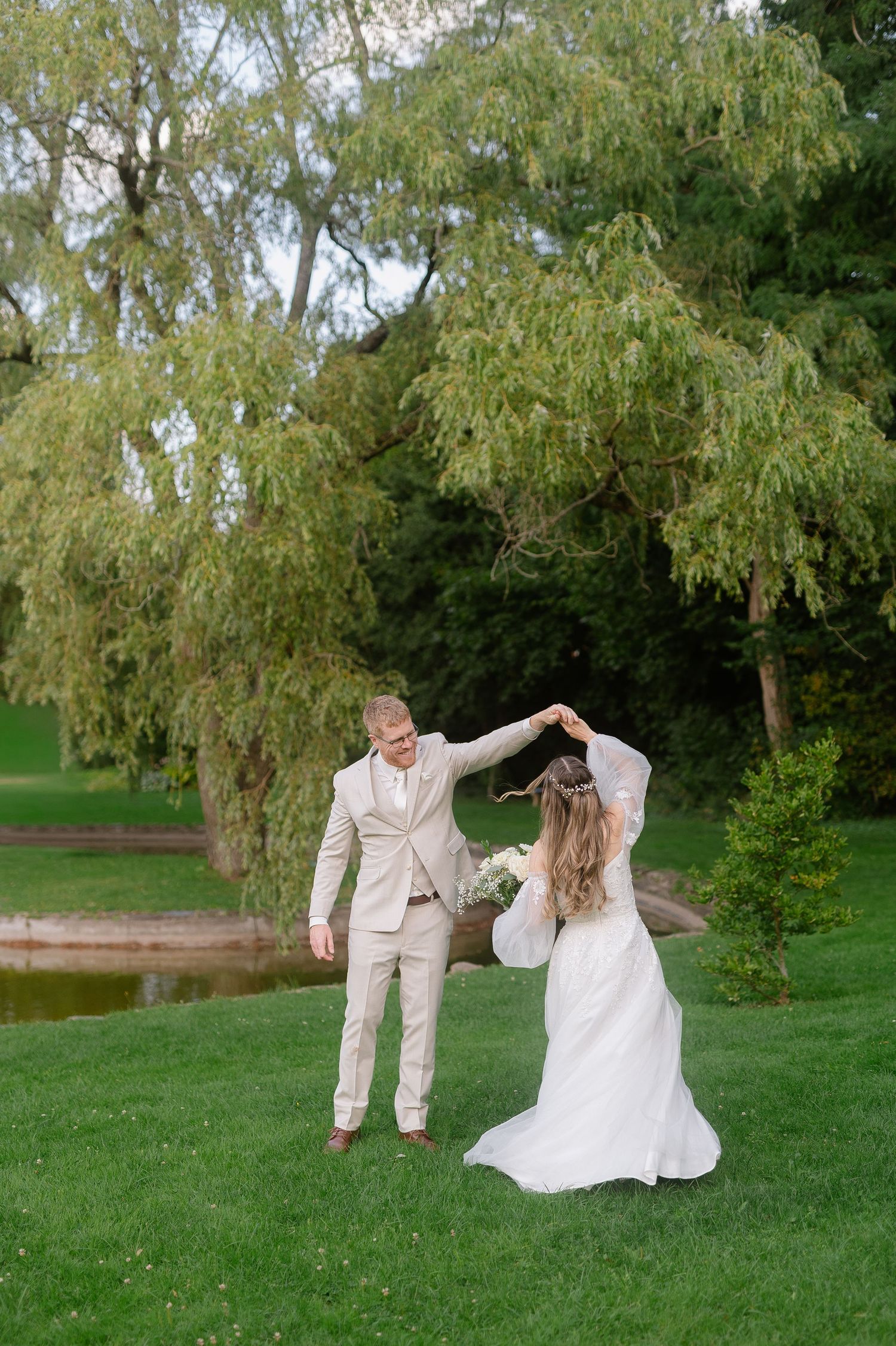A couple dances playfully on lush green grass beneath a weeping willow tree.