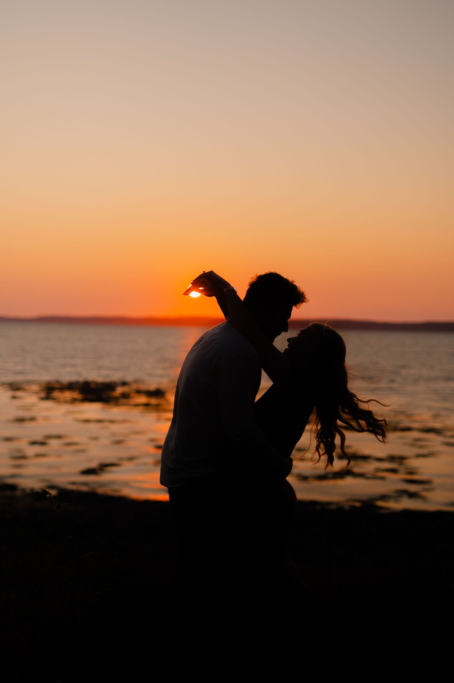 Silhouettes of a couple against a dramatic orange sunset over the ocean.