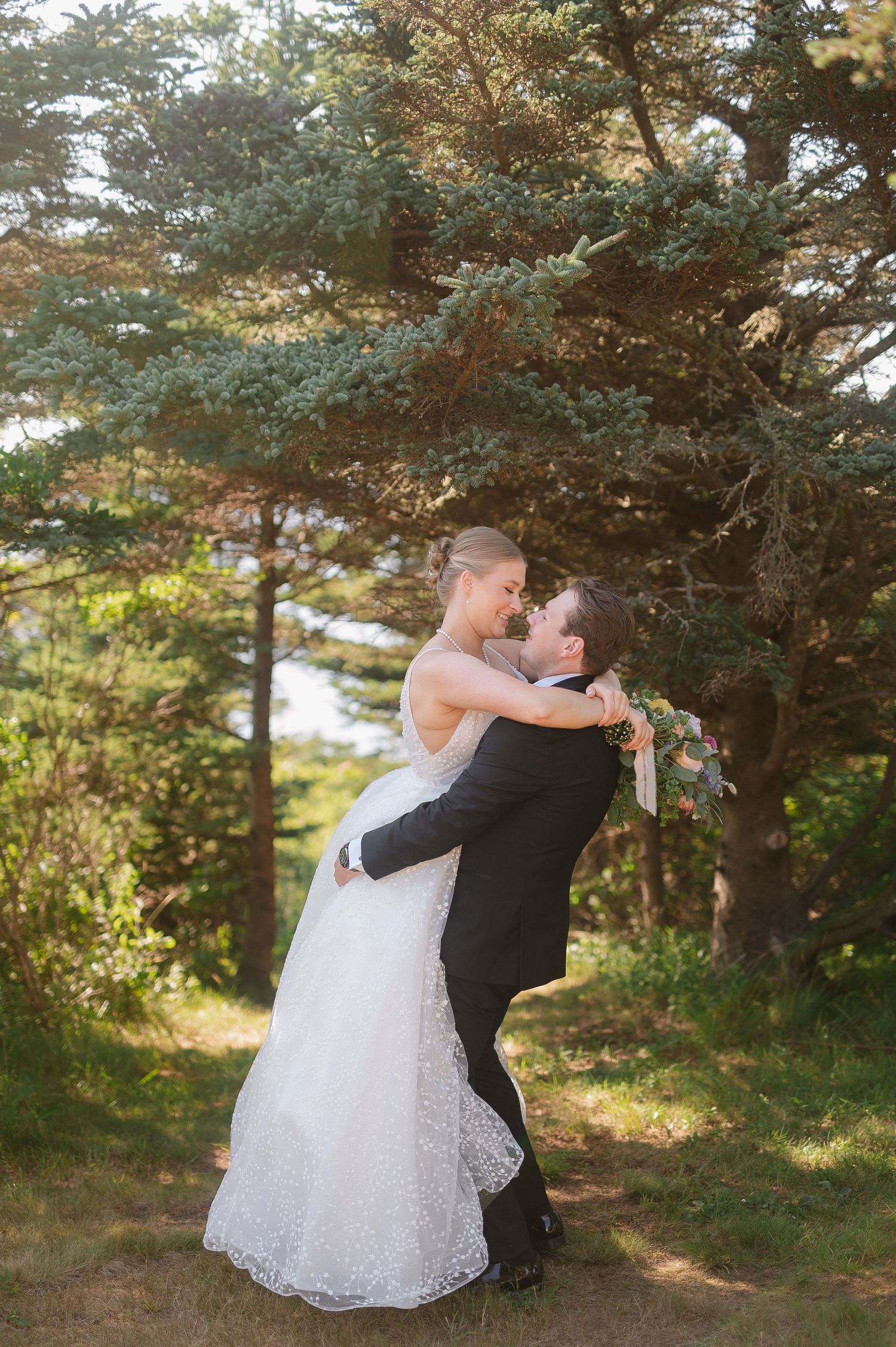 Romantic wedding couple embraces among evergreen trees in natural outdoor setting.