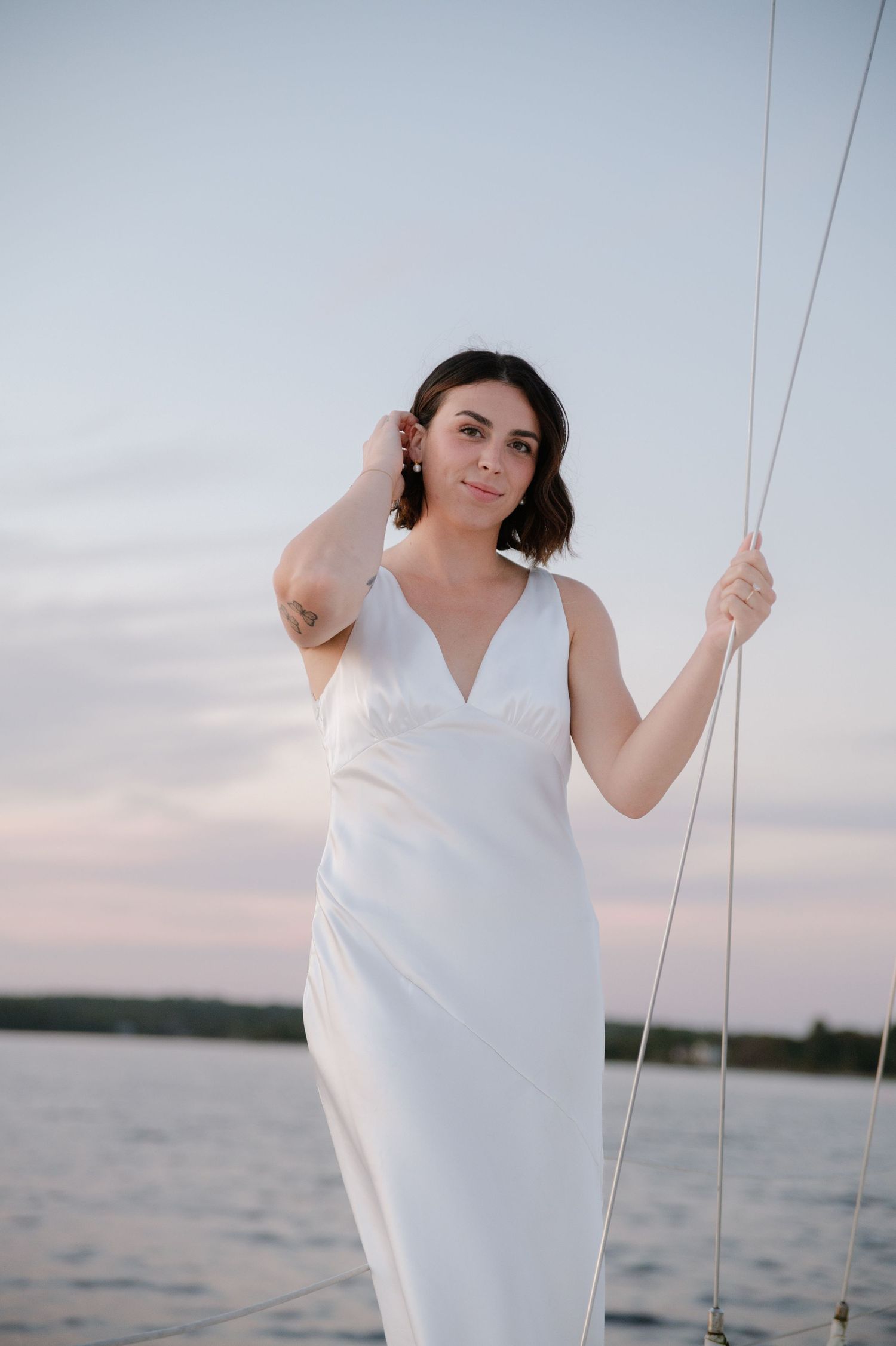 Woman in sleeveless white dress poses gracefully on sailboat during sunset.
