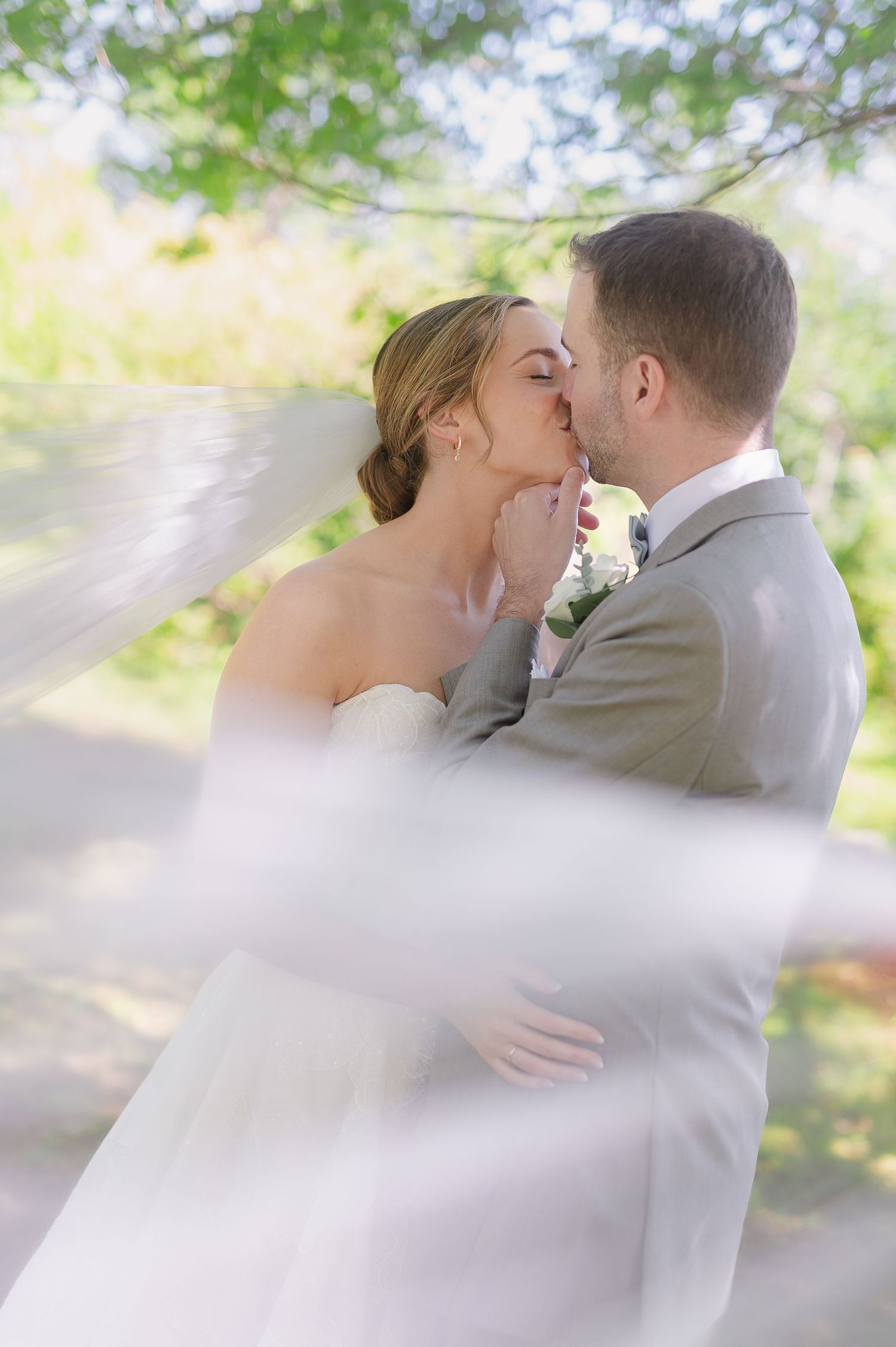 Romantic wedding kiss captured through flowing white veil in garden setting.