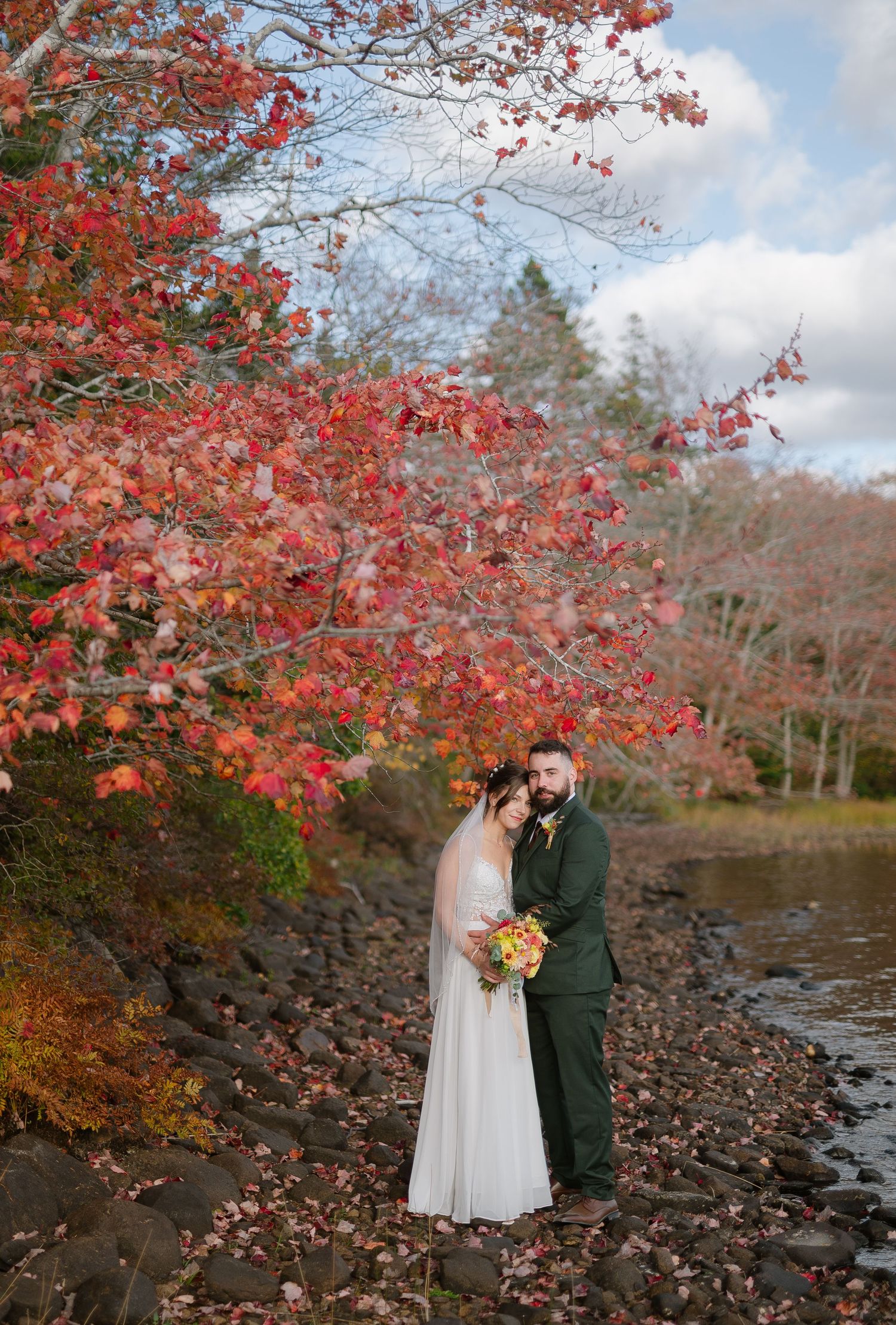 A couple stands together by a lake surrounded by vibrant red autumn foliage.