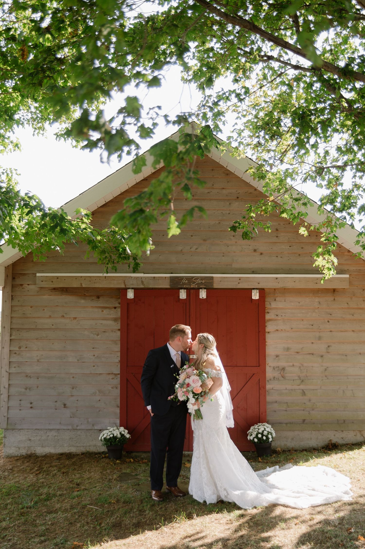 Wedding couple poses against rustic red barn doors in natural outdoor setting.