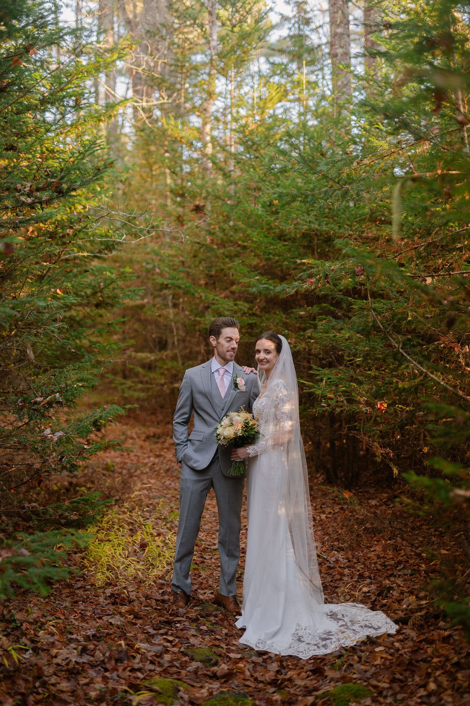 A wedding couple walks through a sunlit autumn forest path surrounded by golden foliage.