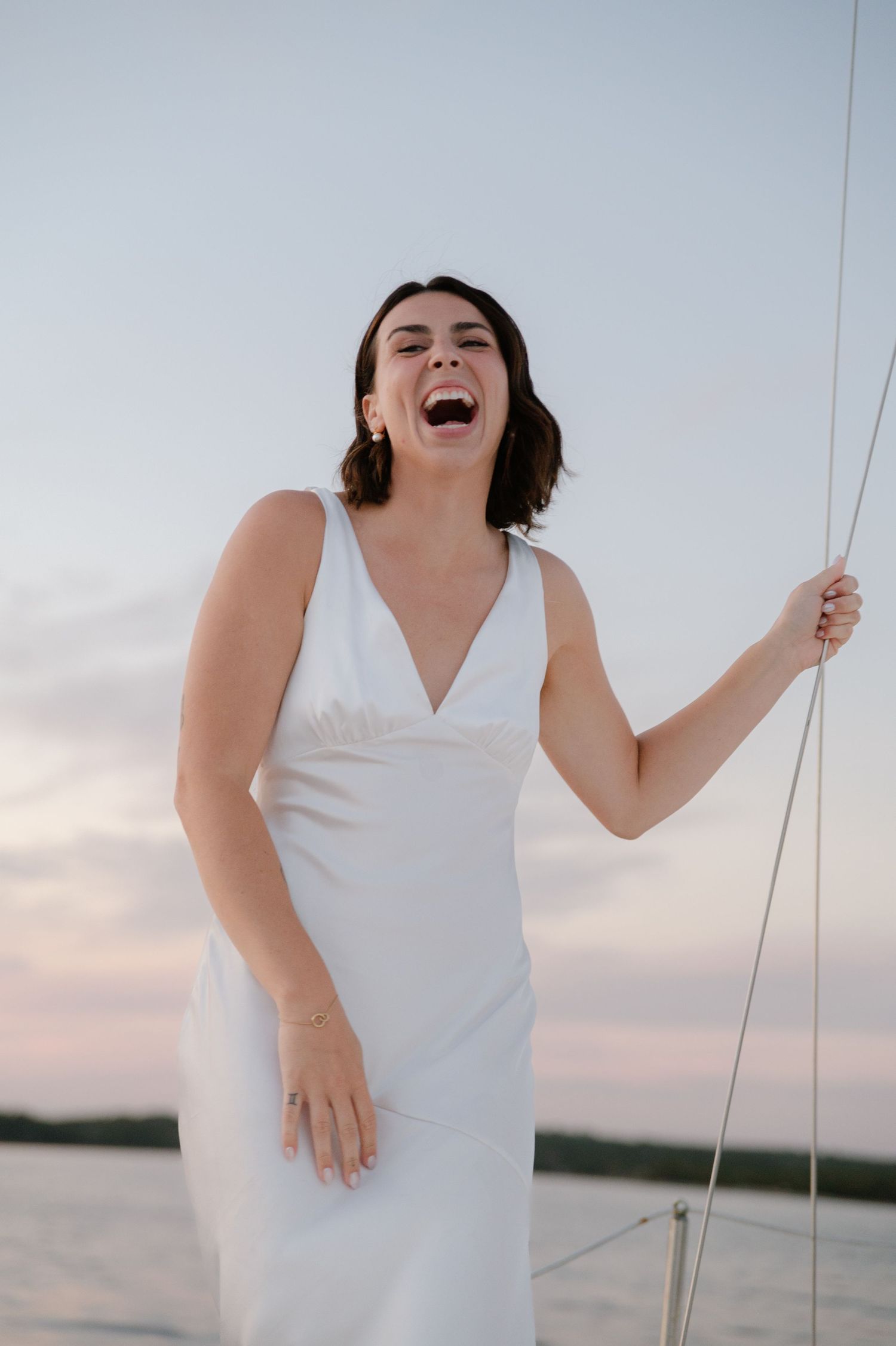 Person in white dress laughs joyfully while holding sailing ropes against sunset sky.