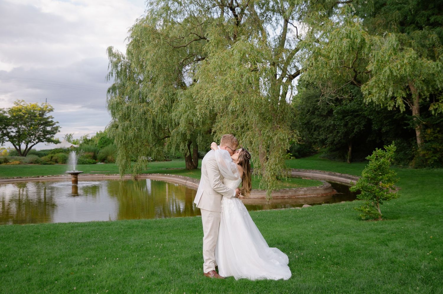 A couple shares a romantic moment under a weeping willow tree beside a tranquil pond at a scenic outdoor venue.