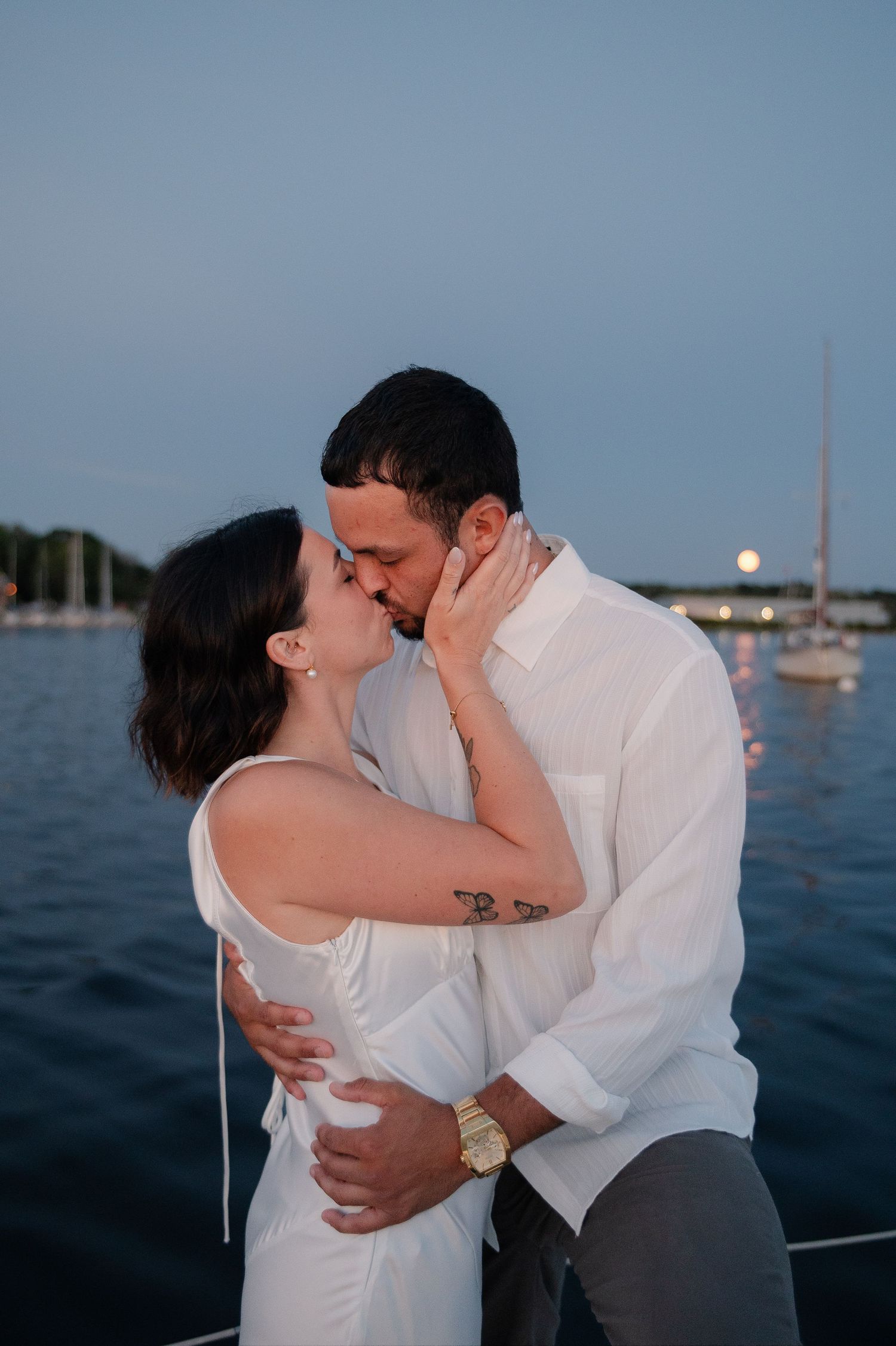 Romantic evening photoshoot of a couple embracing on a boat with sailboats in the background.