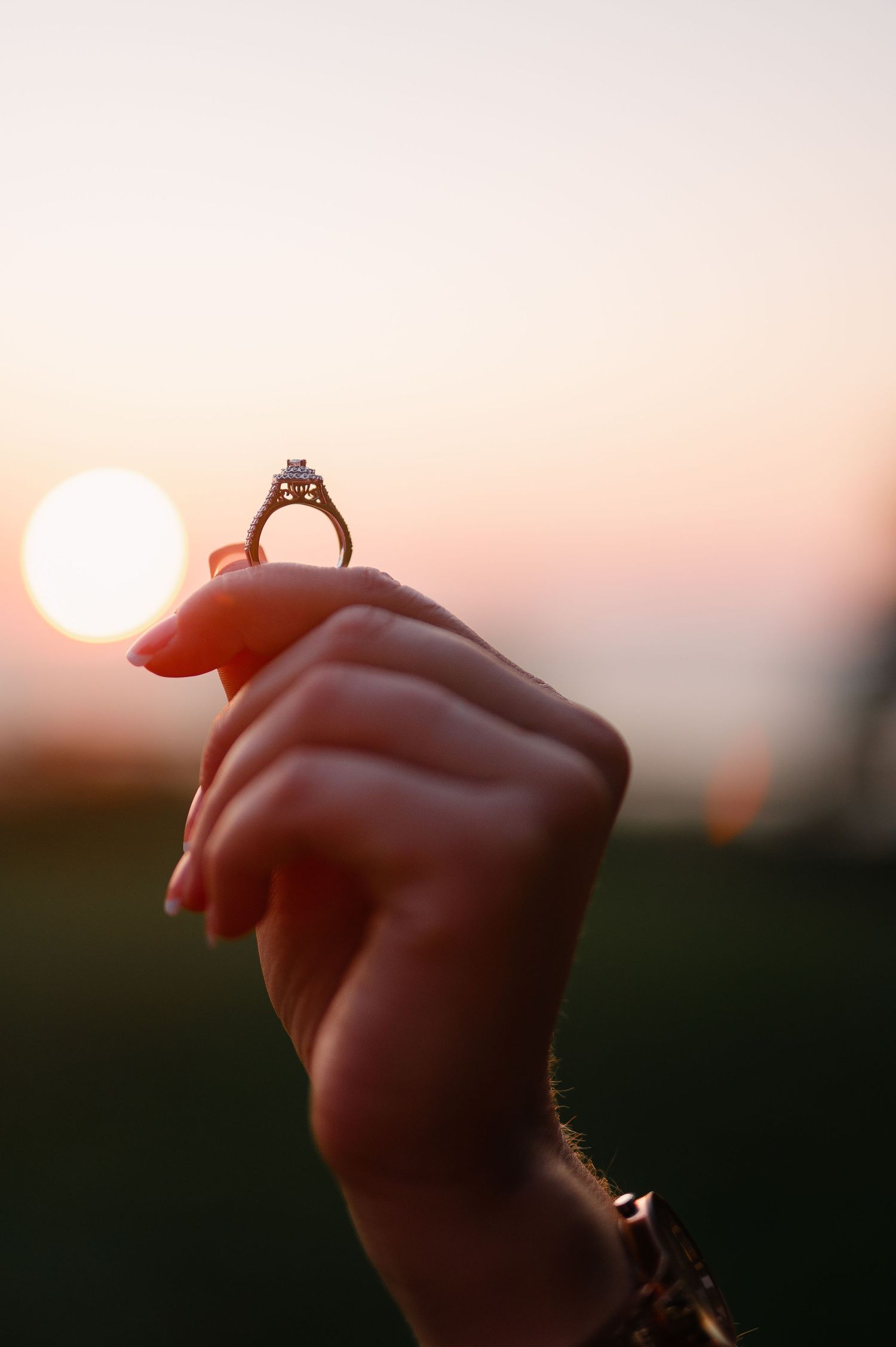A hand holds up an engagement ring against the setting sun.