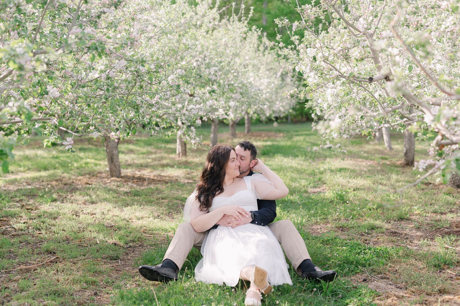 Couple sharing romantic moments on grass beneath flowering trees.