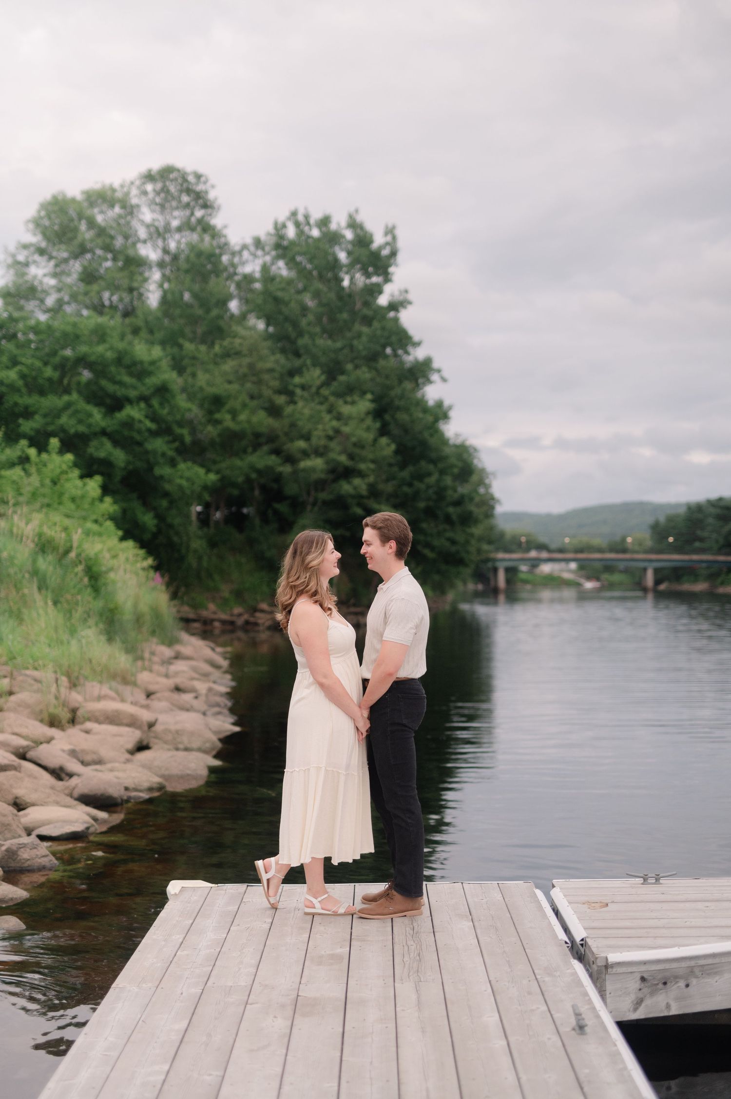 A couple sits together on a wooden dock overlooking a serene river and bridge.
