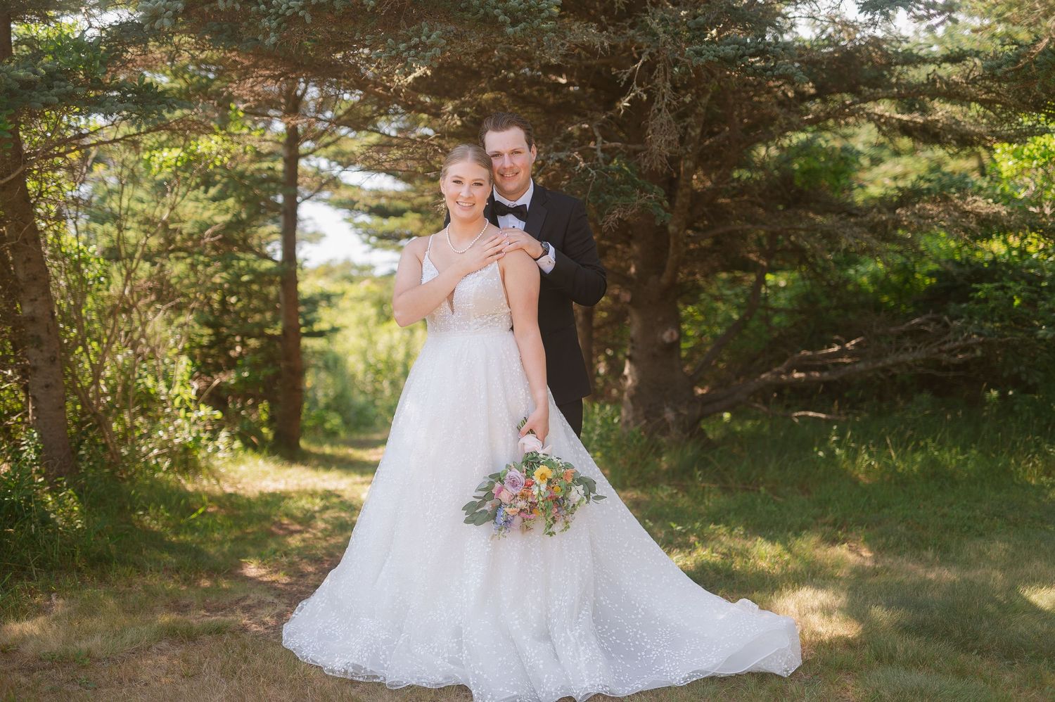 A bride in a white lace gown poses outdoors in a sunlit garden setting with natural greenery in the background.