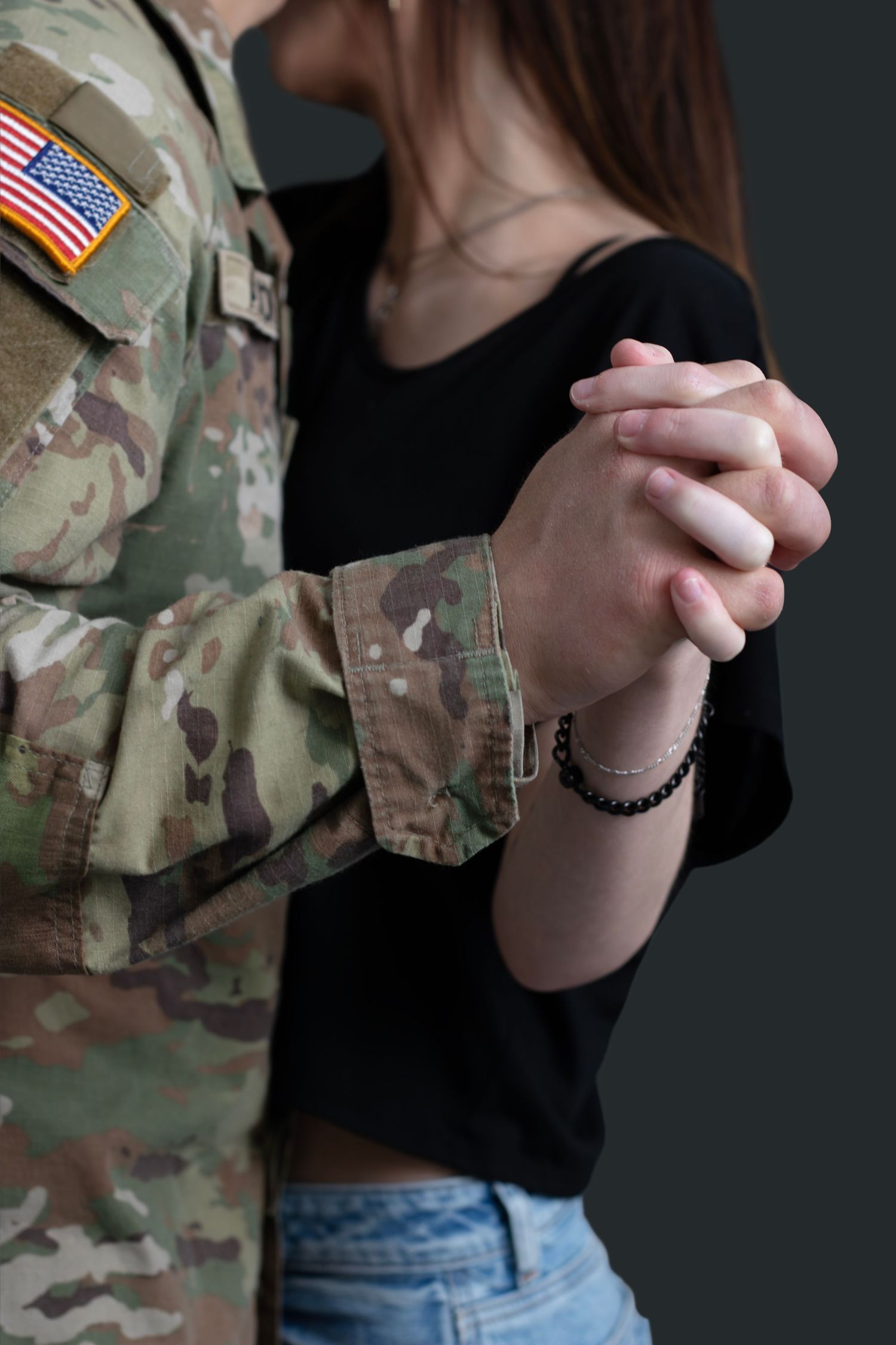 A soldier in camouflage uniform with US flag patch holds hands with woman wearing a black top and jeans at a professional studio in SD.