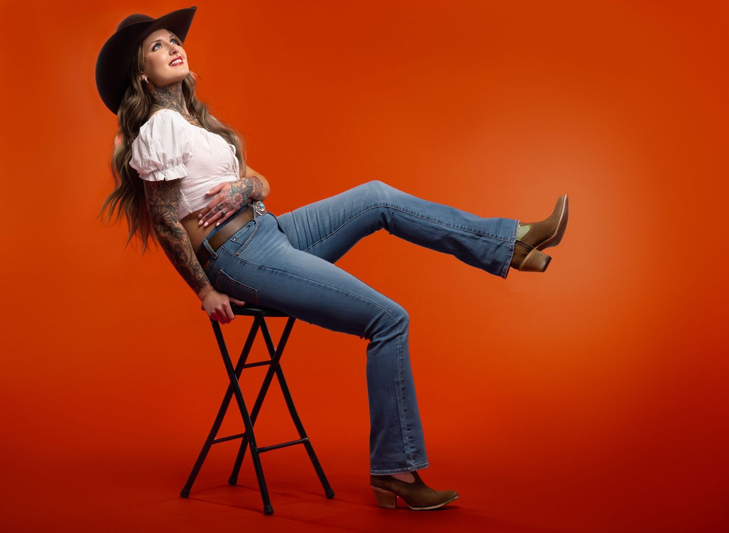 Cowgirl fashion portrait of tattooed woman wearing cowboy hat, white blouse, blue jeans, and boots posing on stool with orange background by