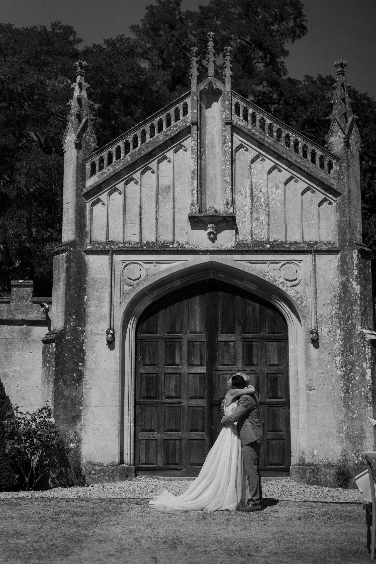 Bride and groom embracing before ornate Gothic arched wooden doors of a historic stone manor in black and white.