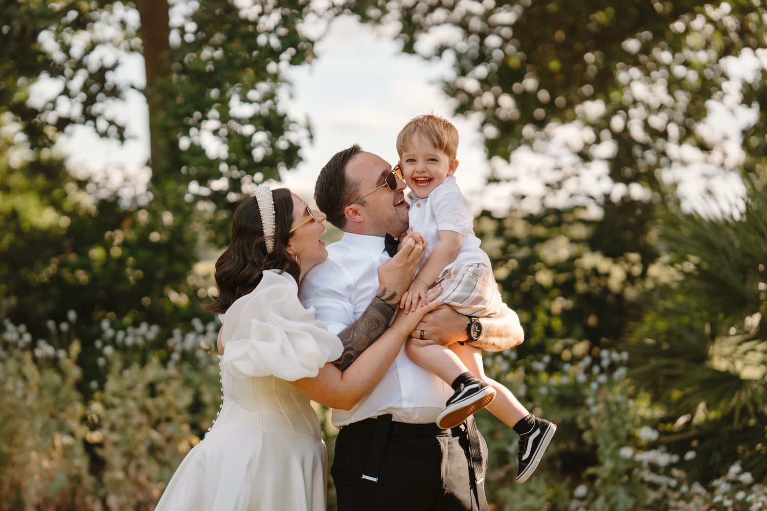 Bride and groom in wedding attire joyfully kissing their smiling toddler outdoors among lush green trees.