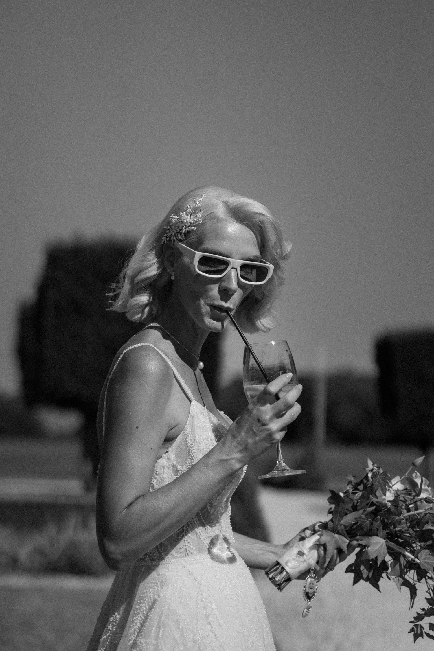 Black and white photo of a bride in white dress and sunglasses sipping champagne while holding a bouquet outdoors.
