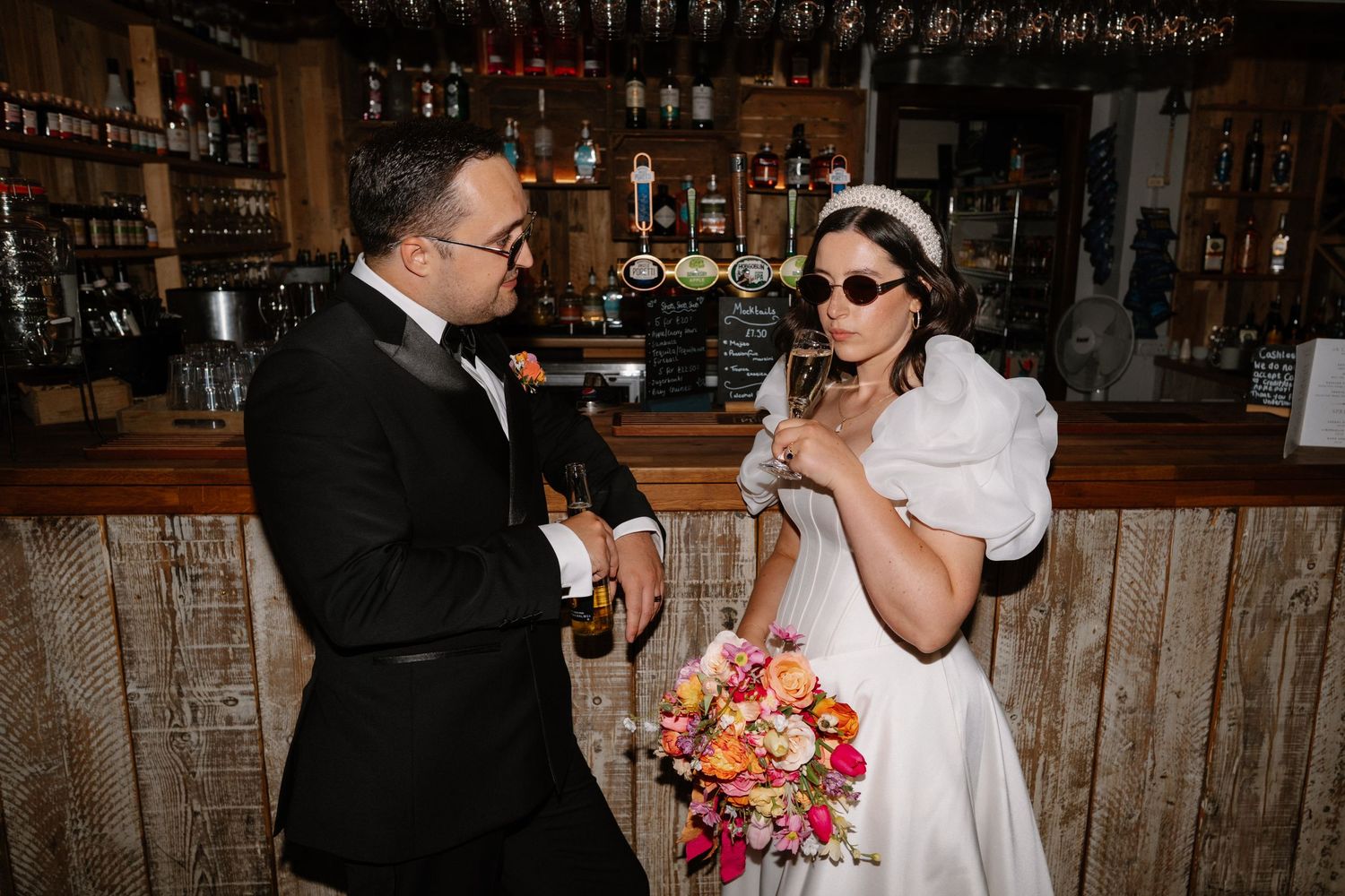 Bride in white gown and sunglasses with groom in tuxedo sharing drinks at a rustic wooden bar.