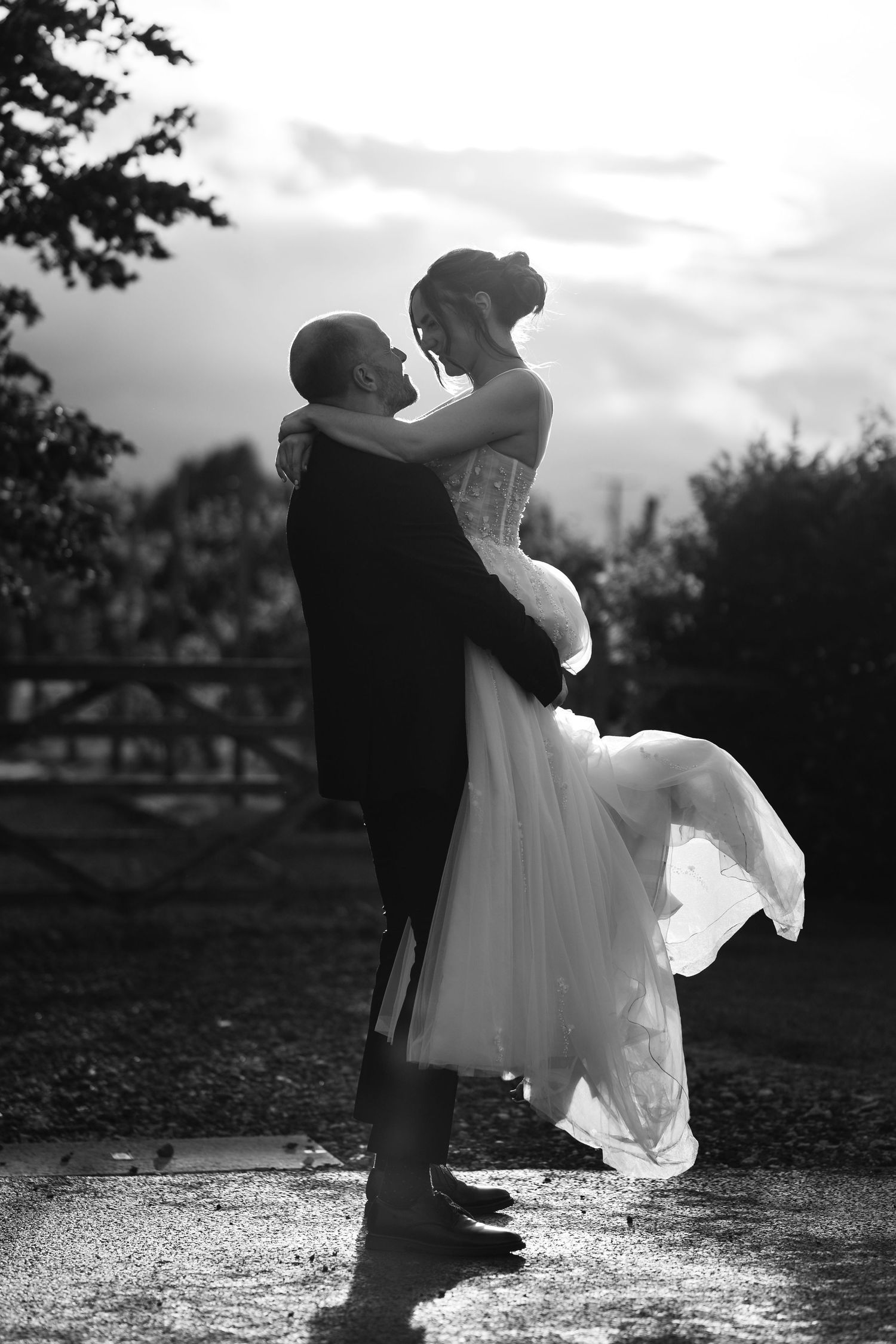 Black and white photo of groom lifting bride in wedding dress outdoors, romantic sunset silhouette portrait.