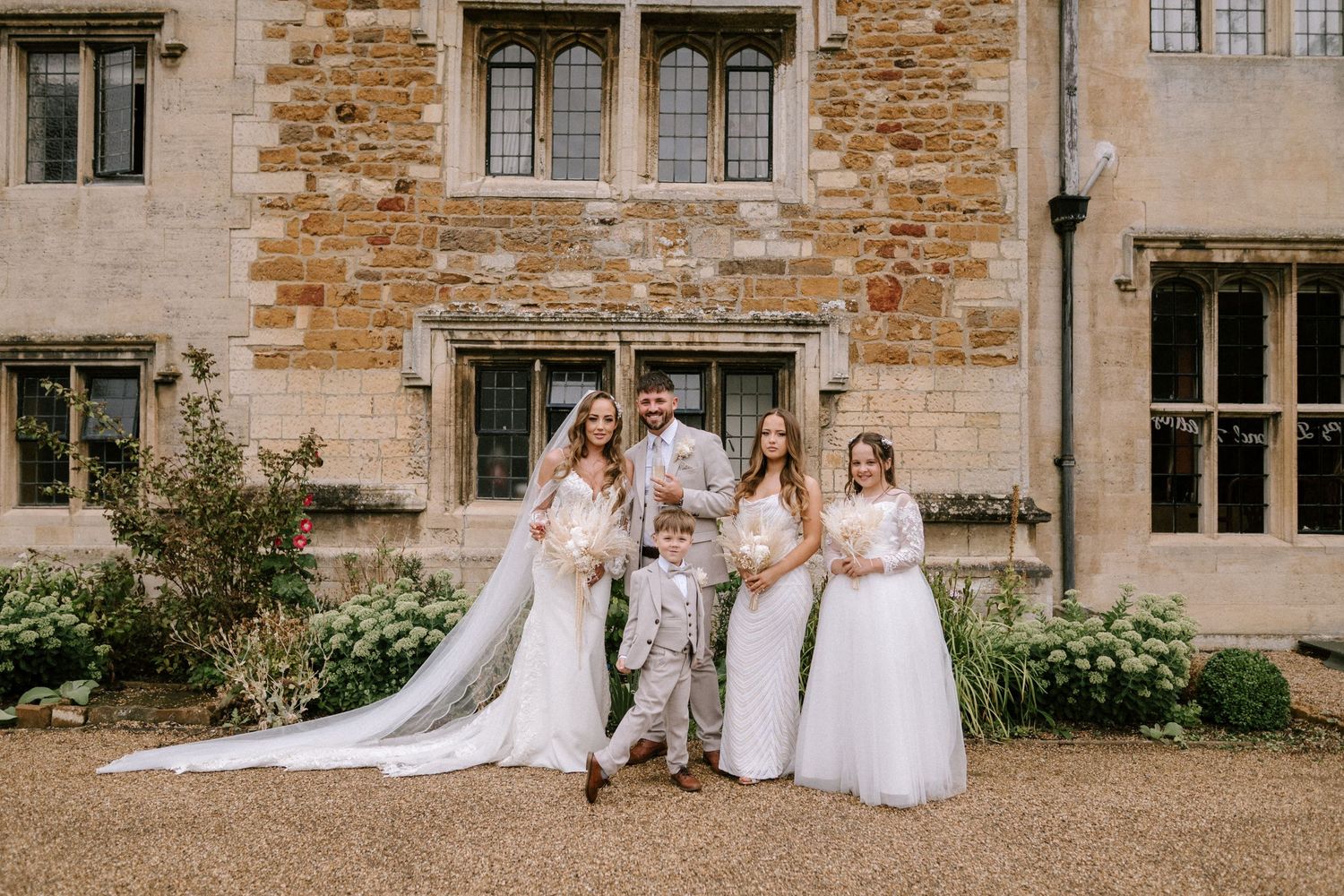 A group in formal wedding attire poses together outside a historic stone manor house building.