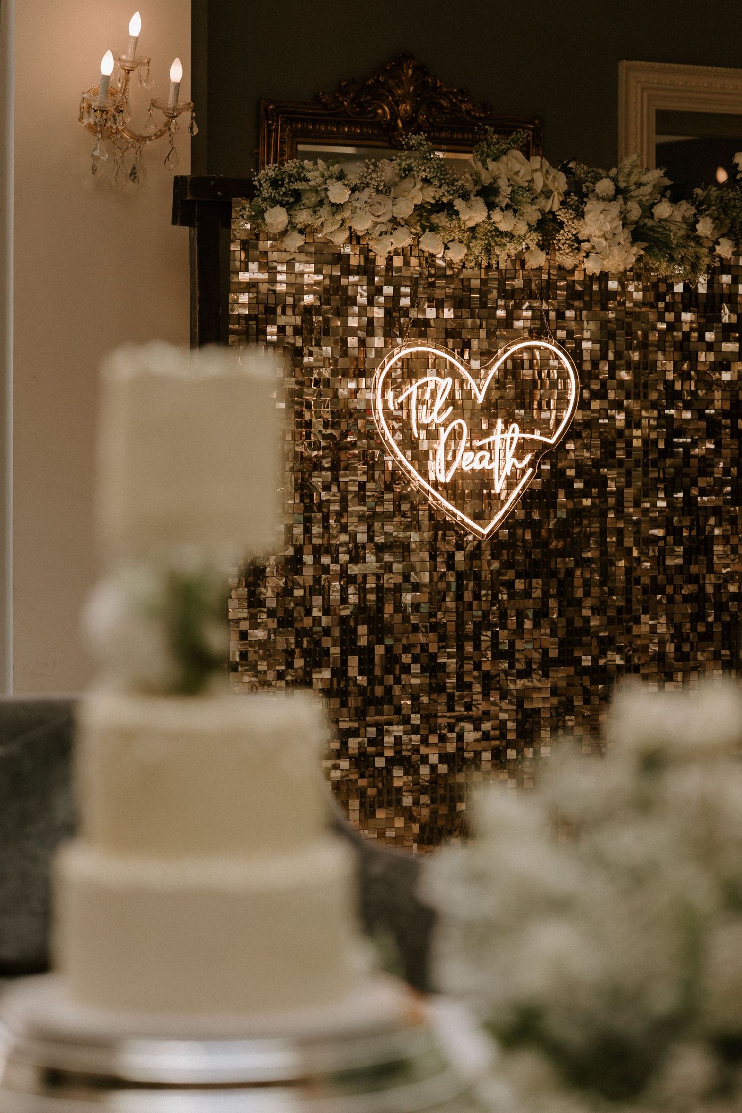 Wedding cake display with illuminated heart neon sign and sparkly sequined backdrop.