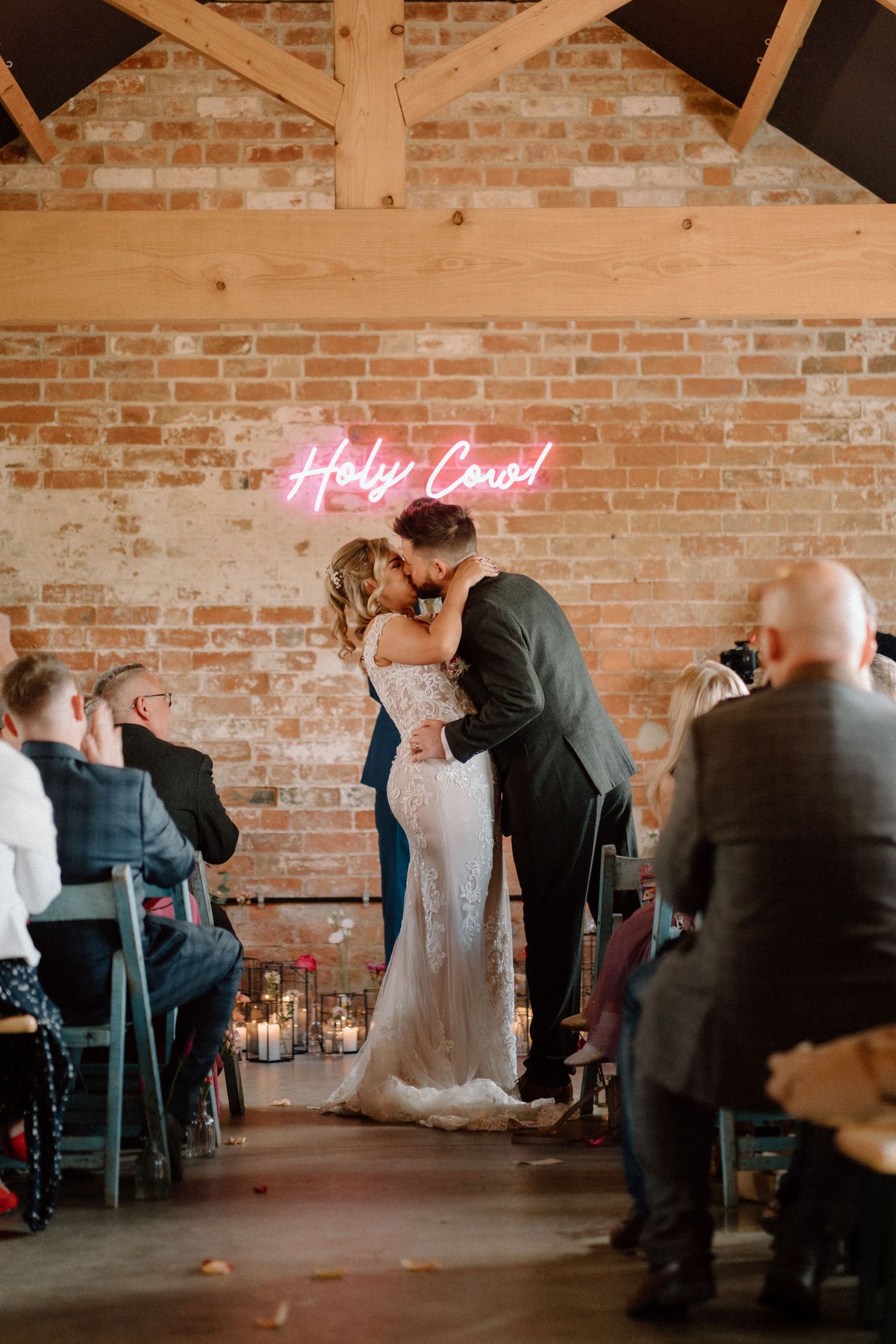 A wedding ceremony in a rustic brick venue with a pink neon 'Holy Cow' sign glowing above at The Old Cow Shed.