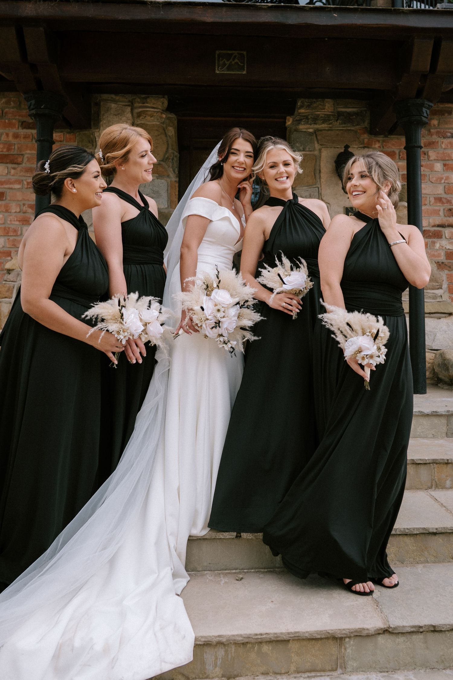 Bridal party on stone steps with bride in white gown and bridesmaids in elegant black dresses holding bouquets.
