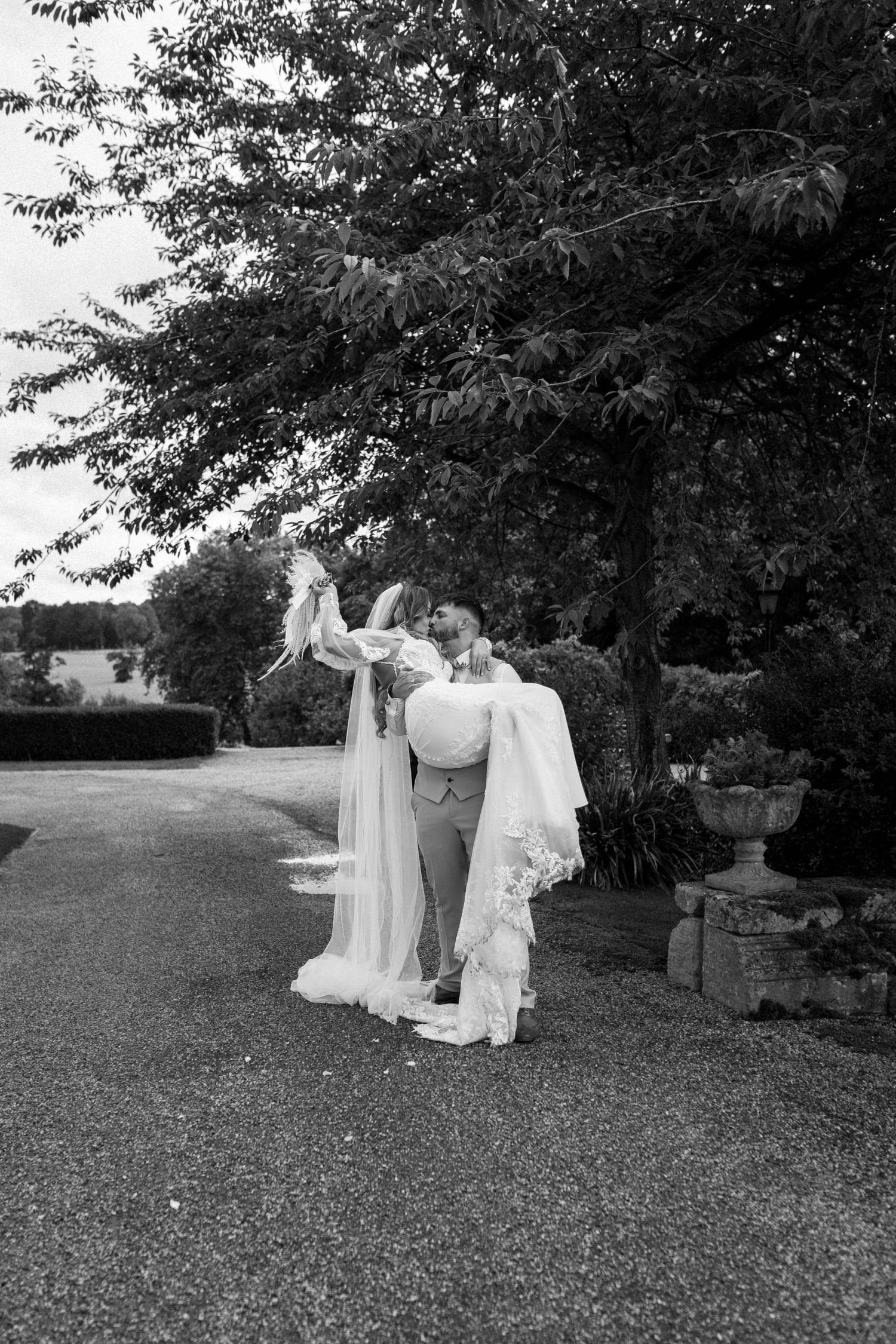 Two people adjust a flowing wedding veil in a beautiful garden setting with classic stone features.