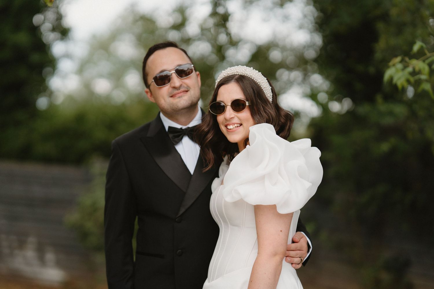 A bride and groom in sunglasses pose outdoors, she in a white ruffled dress and he in a black tuxedo.