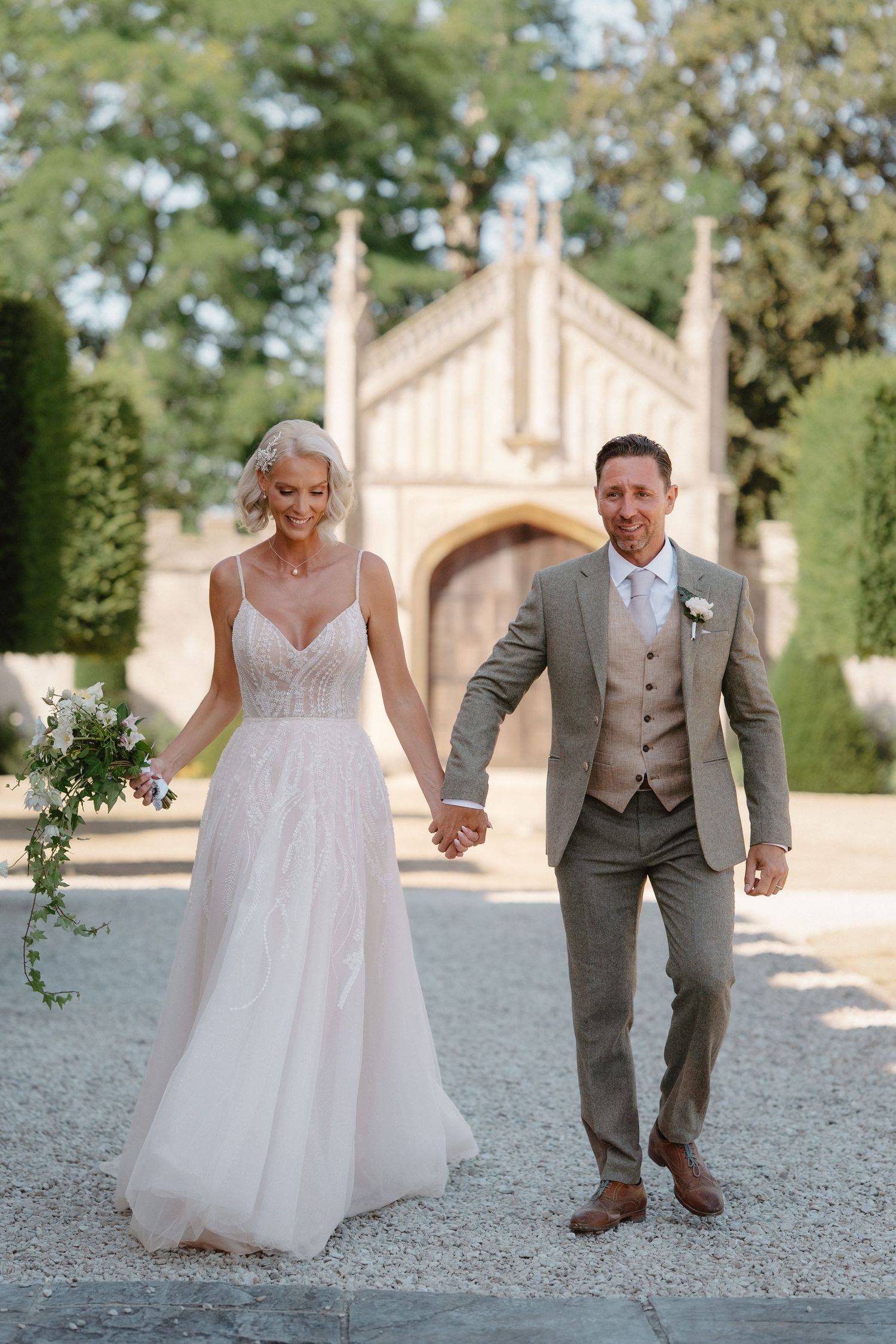 Smiling bride and groom holding hands walking outdoors with Gothic stone chapel and lush trees in background.