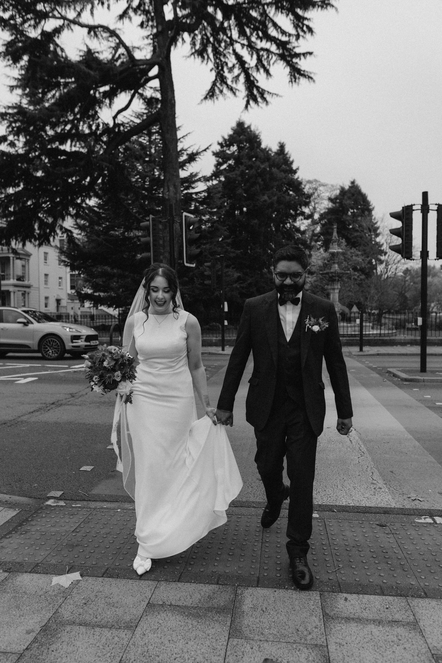 A couple walks together on an urban sidewalk in wedding attire, captured in black and white at The Glasshouse in Leamington Spa.