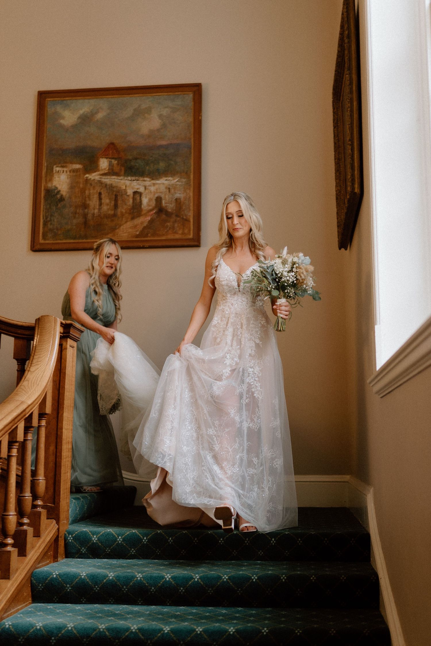 Bride descending carpeted stairs in a lace gown while bridesmaid helps with the dress in Warwickshire.