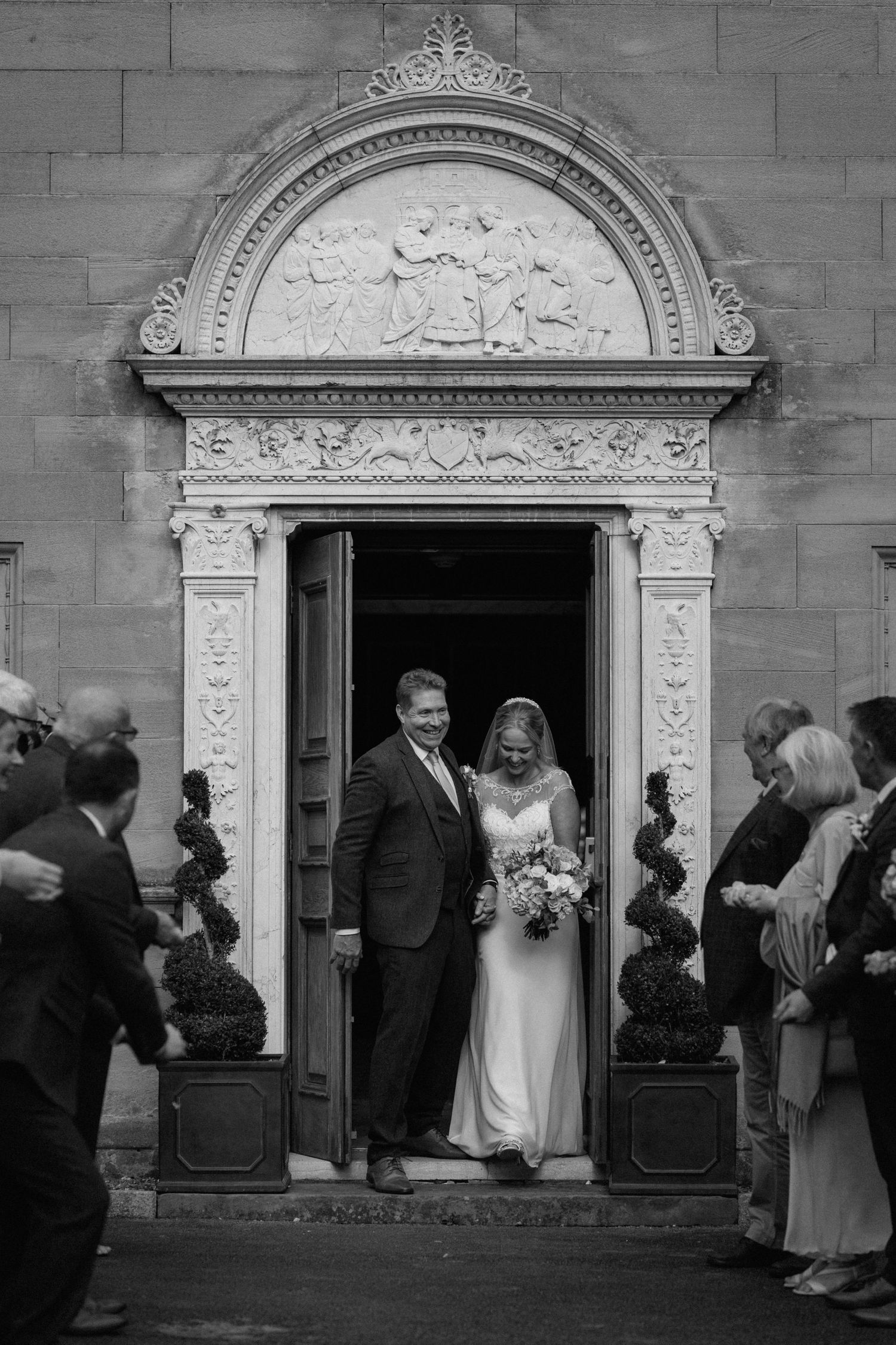 A couple exits through an ornate stone doorway with decorative carved archway after their wedding ceremony at Bourton Hall.