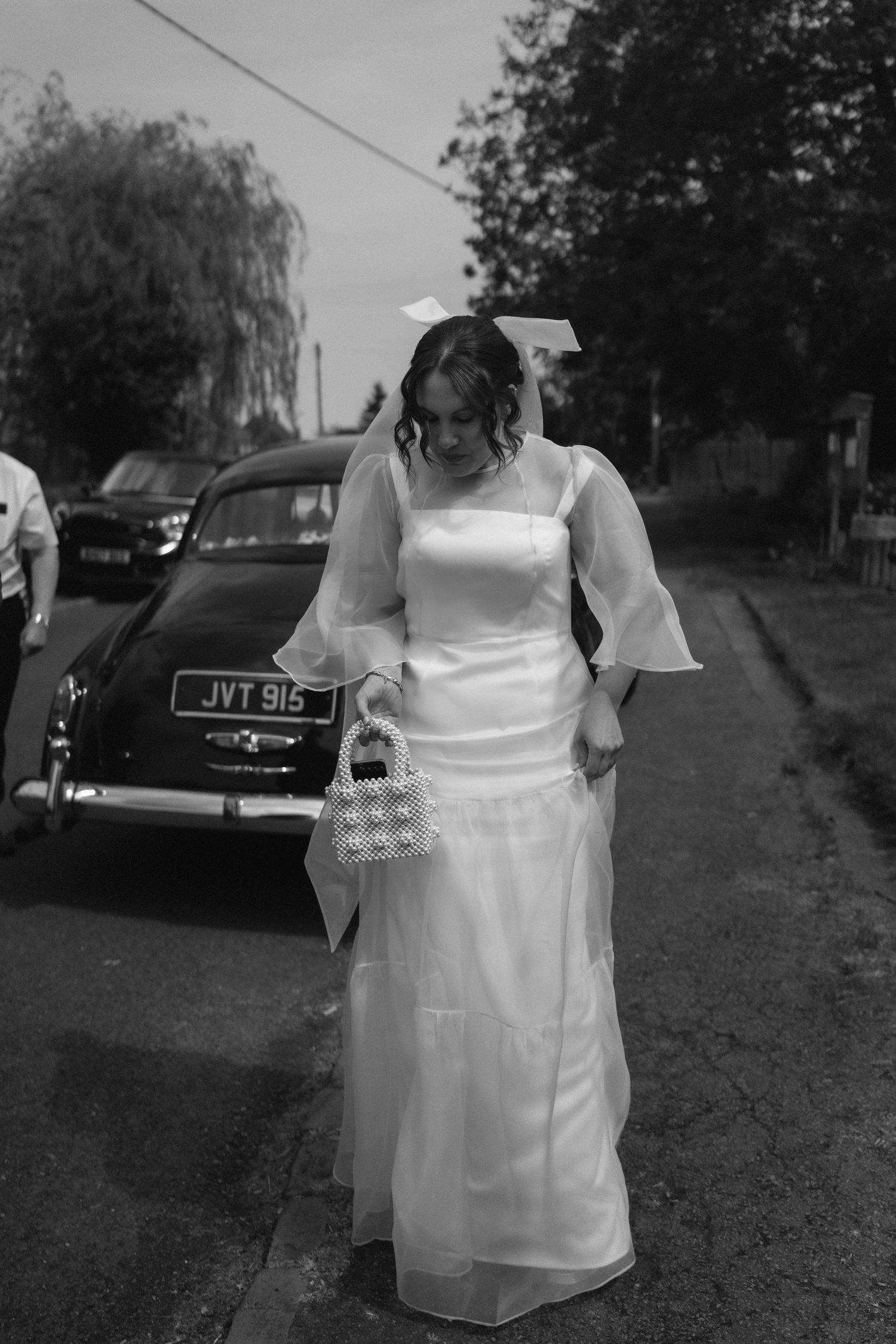 A vintage-style black and white photograph of a wedding dress with bell sleeves and a long veil next to a classic car in Warwickshire.