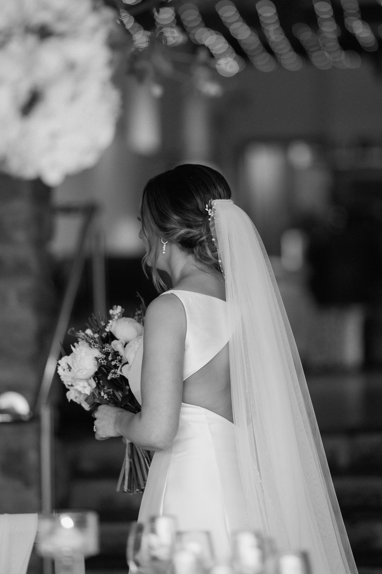 A bride in a white wedding dress with an open back detail holds her bouquet while wearing a long flowing veil at Dodford Manor.