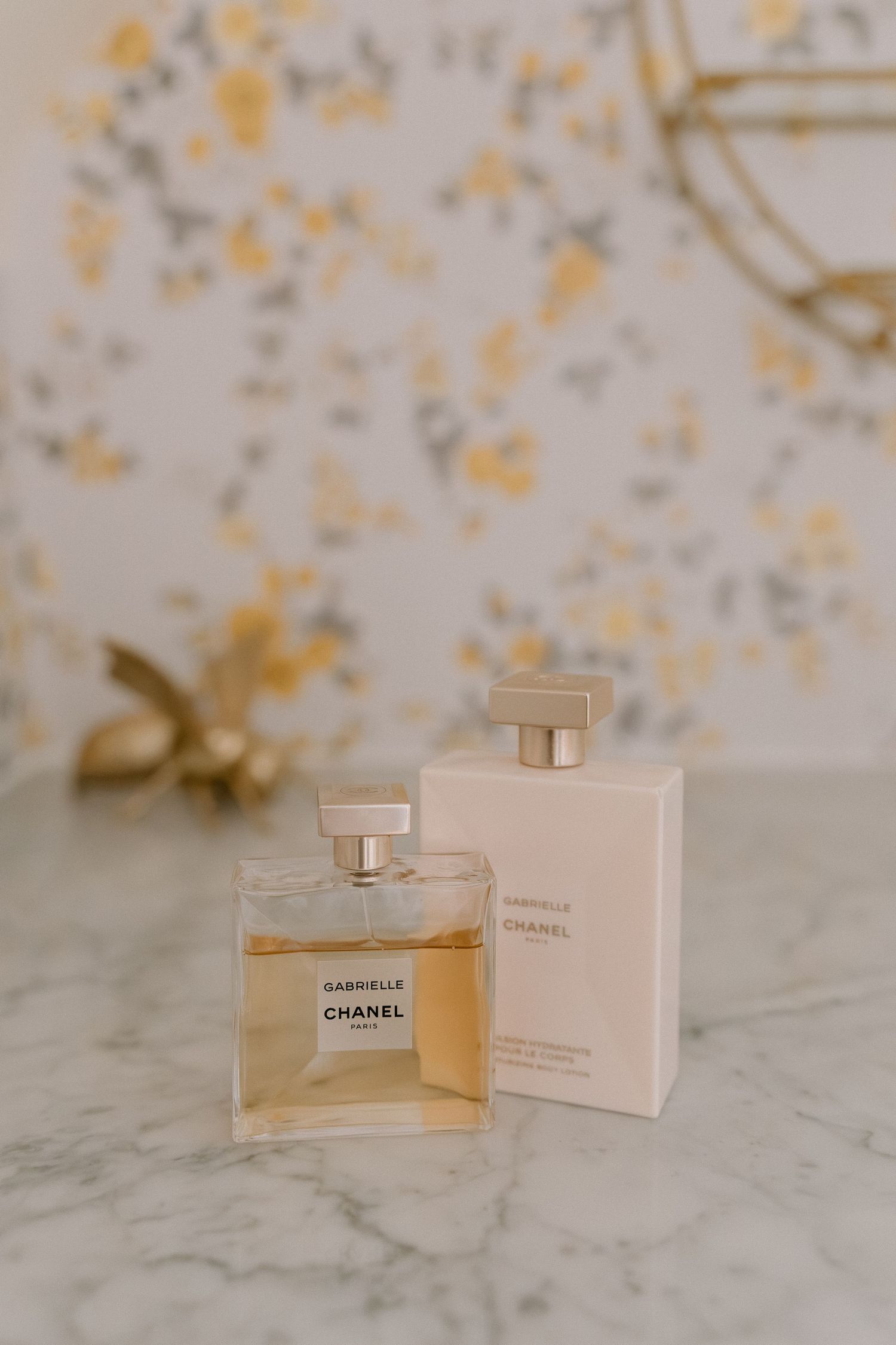 Chanel Gabrielle perfume bottle and box on marble surface with floral wallpaper background.