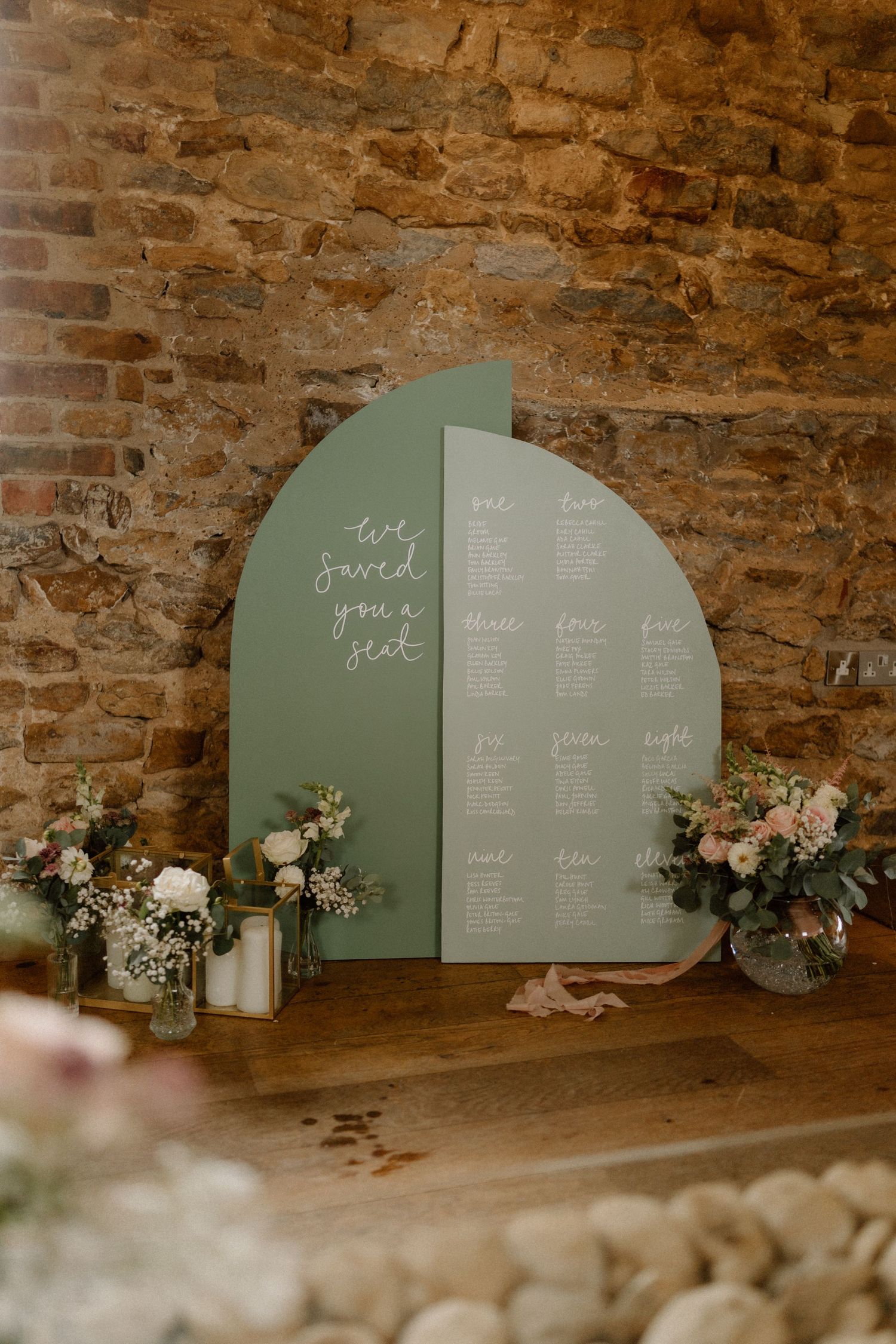 Sage green semicircle seating chart display with floral arrangements against brick wallat Dodford Manor.