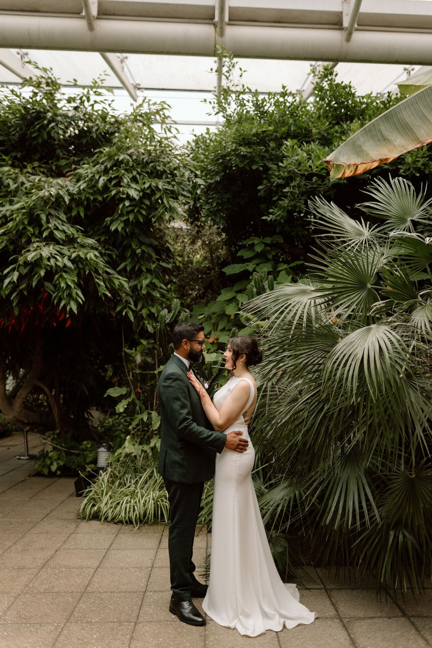 Newlyweds share an intimate moment in a greenhouse setting surrounded by lush palms and greenery at The Glasshouse in Leamington Spa.