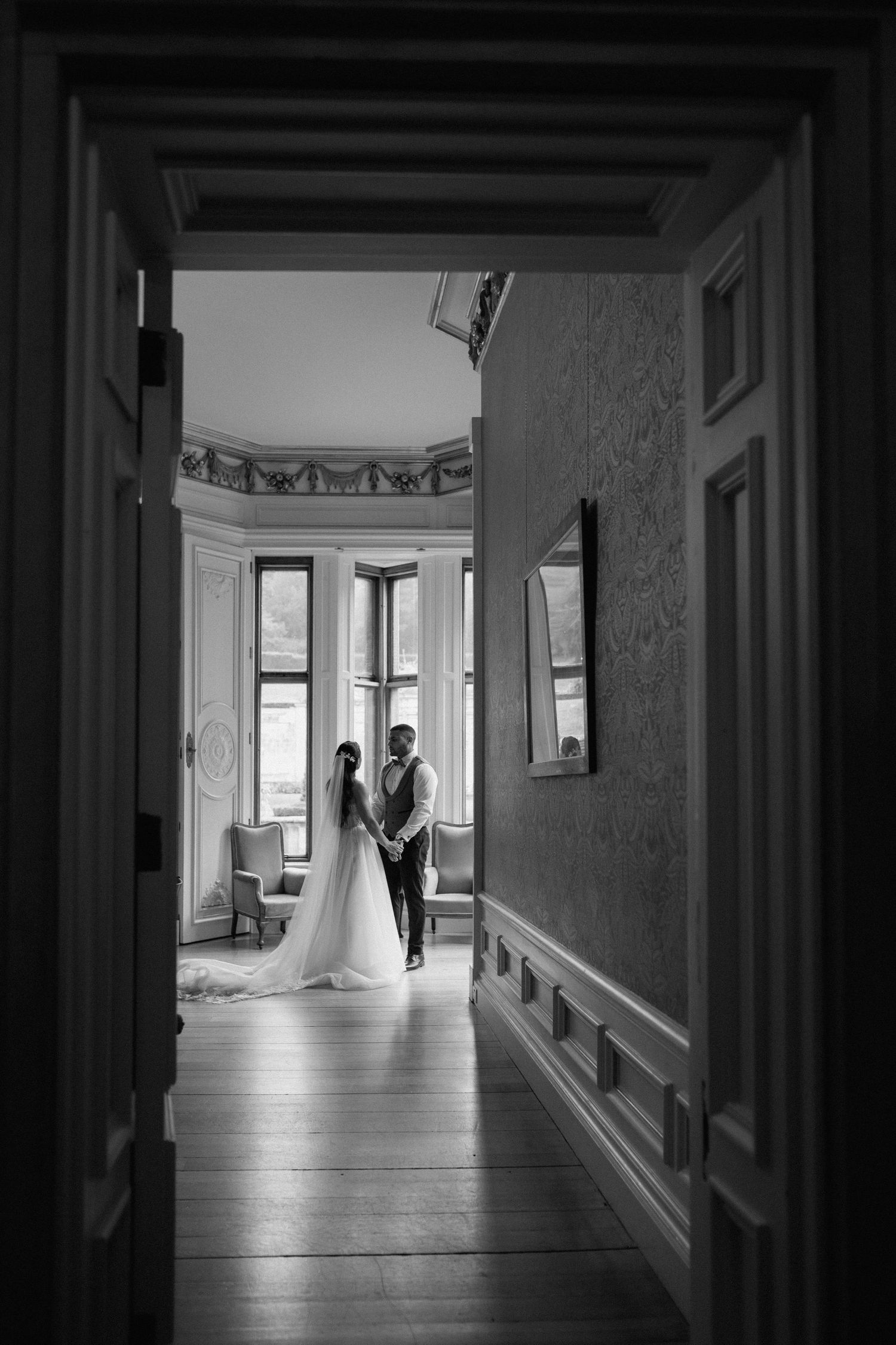 A black and white photo through a doorway showing a couple in a sunlit room with bay windows at Harlaxton Manor.