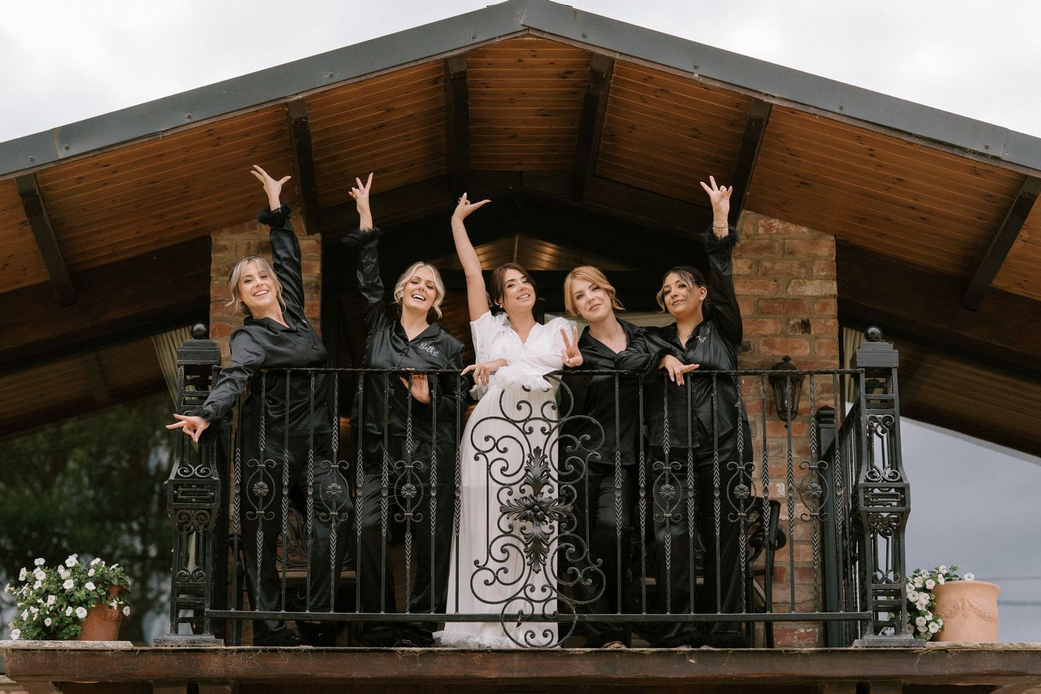 Wedding party members gather on a rustic wood balcony with decorative black iron railings for a formal event photo.
