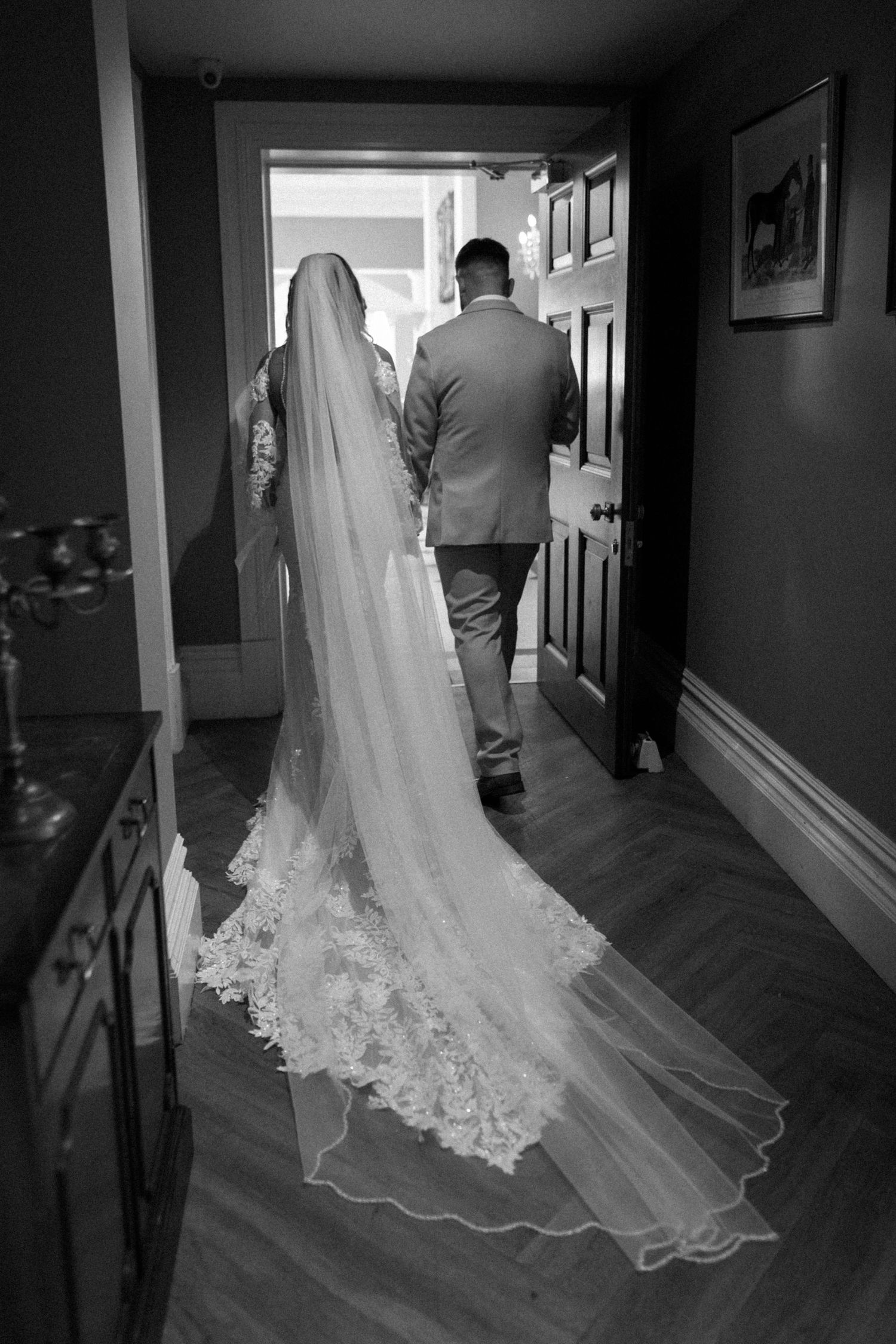 A trailing lace wedding veil follows behind as they walk through a doorway.