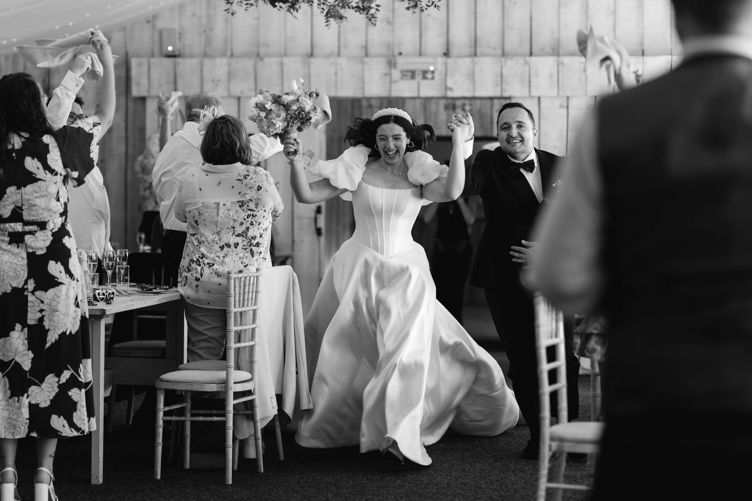 Black and white photo of a bride in a white gown entering a wedding reception, waving joyfully to guests.