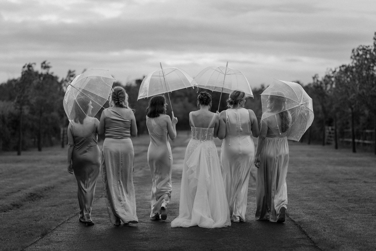 Bride and bridesmaids holding clear umbrellas stand together outdoors on a cloudy wedding day.