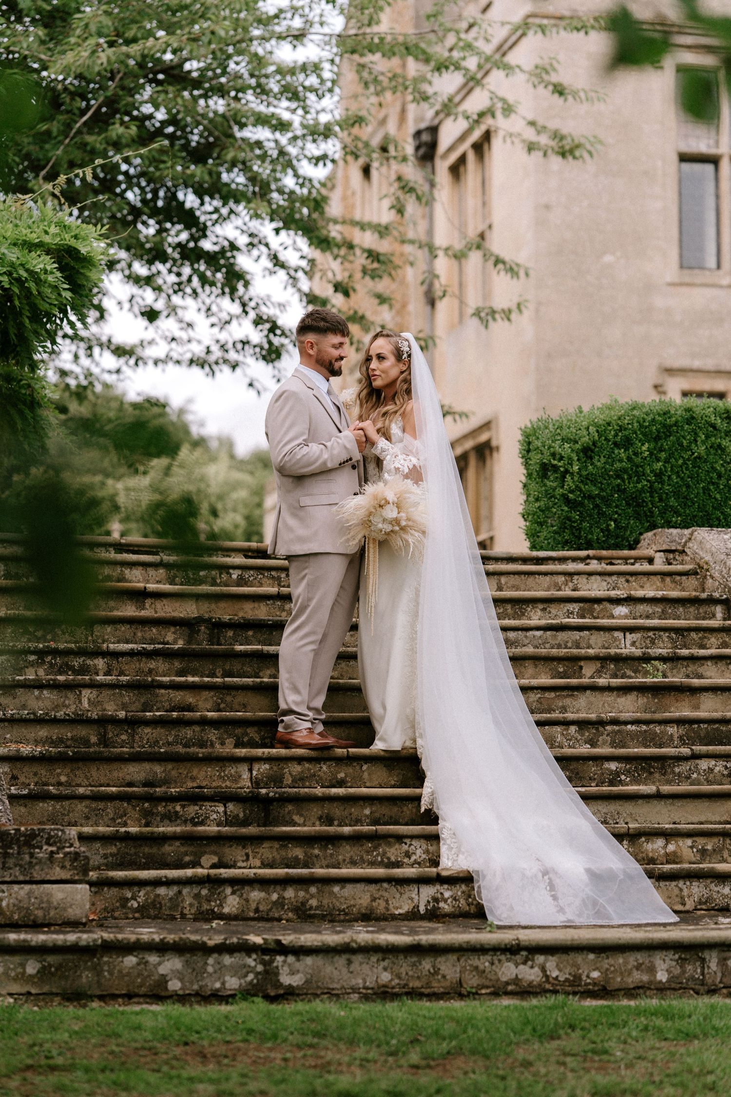 Wedding couple poses on historic stone staircase surrounded by lush gardens. Wedding photography at Rushton Hall. Northamptonshire Wedding